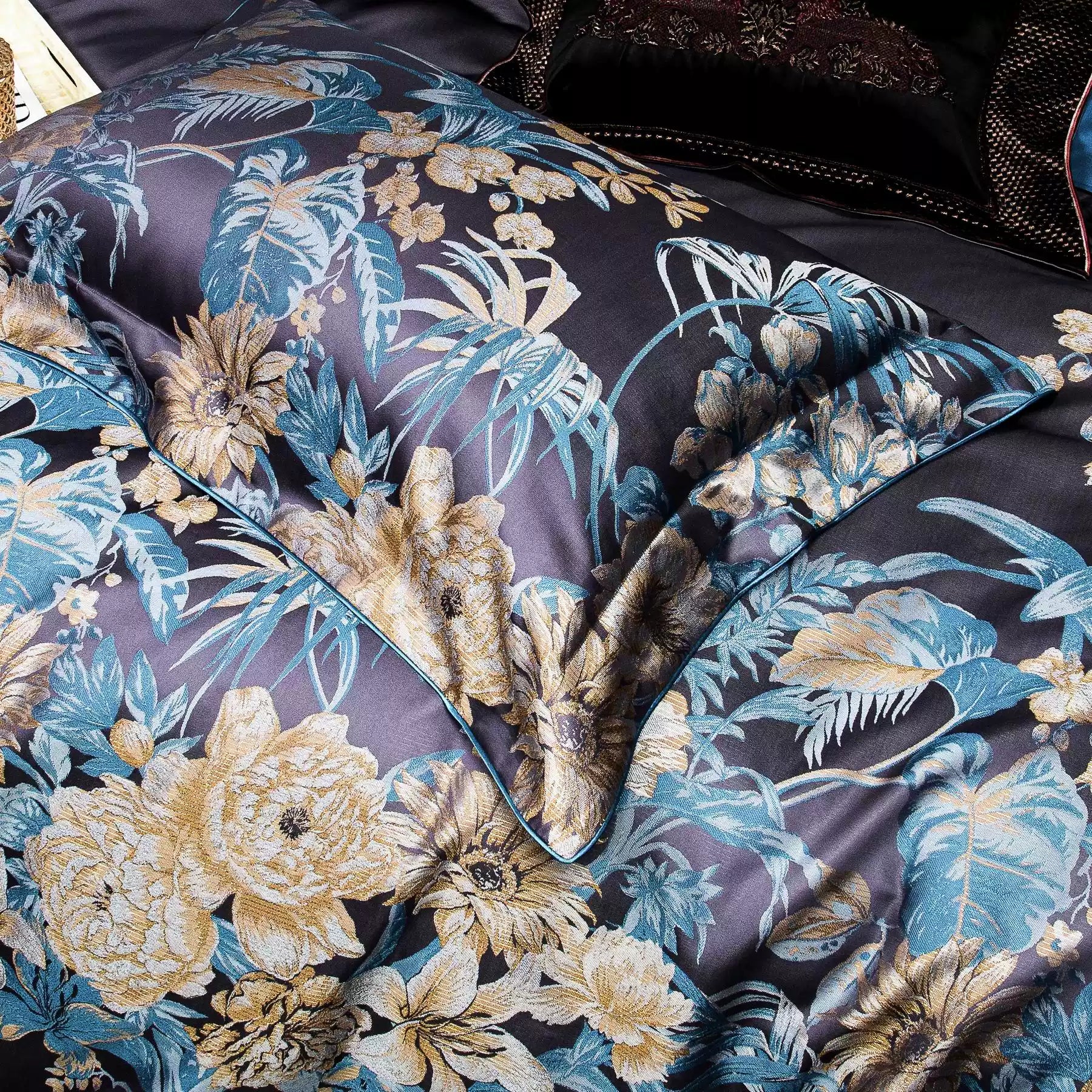 This image showcases a close-up of the pillowcase from the luxurious bedding set. The intricate floral patterns in gold and teal hues stand out beautifully against the deep navy background. The design highlights the rich texture and fine detailing, with the piping adding a sophisticated finishing touch. This bedding exudes elegance and is ideal for a refined and stylish bedroom look.