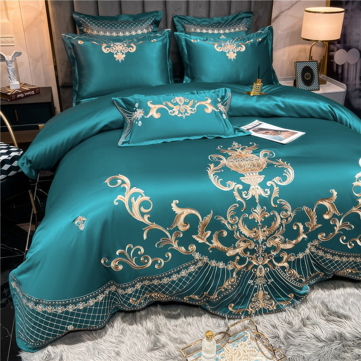 A close-up of a teal satin bedding set adorned with intricate gold embroidery featuring ornate baroque-style scrollwork and floral motifs. The set includes multiple pillows with matching embroidery and a central lumbar pillow, all arranged on the bed. The bedding drapes elegantly, showcasing its sheen and luxurious texture, while a soft gray rug adds a cozy touch to the scene. A tray with pearl accessories and a decorative object enhances the opulent aesthetic of the bedroom.

