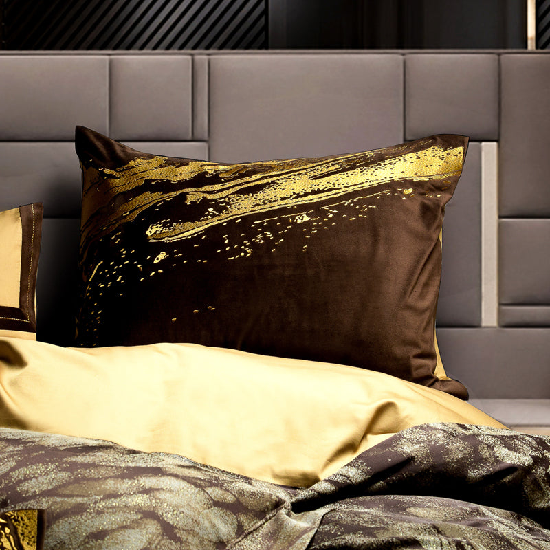 A close-up of a square decorative pillow in deep brown velvet with a striking golden streak design across the top, creating a luxurious and modern aesthetic. The pillow is part of a bedding ensemble, placed against a sophisticated gray, paneled headboard, further accentuating its opulence and contemporary style.