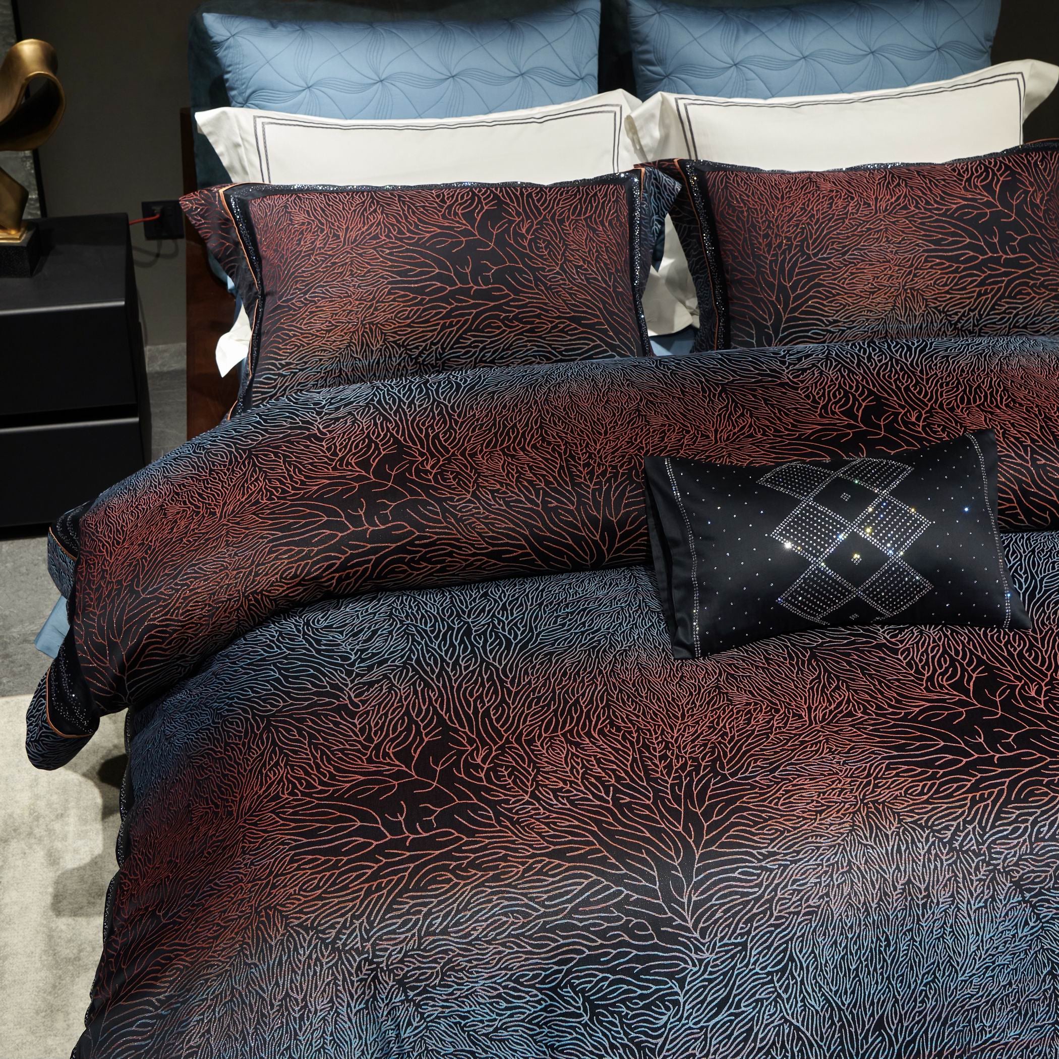 A close-up view of a luxury bedding set featuring a stunning coral-inspired pattern in deep red and navy blue tones. The intricate design is spread across the duvet cover and pillow shams, with a gradient effect transitioning from darker hues to lighter accents. A black decorative pillow with geometric embroidery and sparkling embellishments adds a refined touch, complementing the modern, elegant aesthetic of the bedding.

