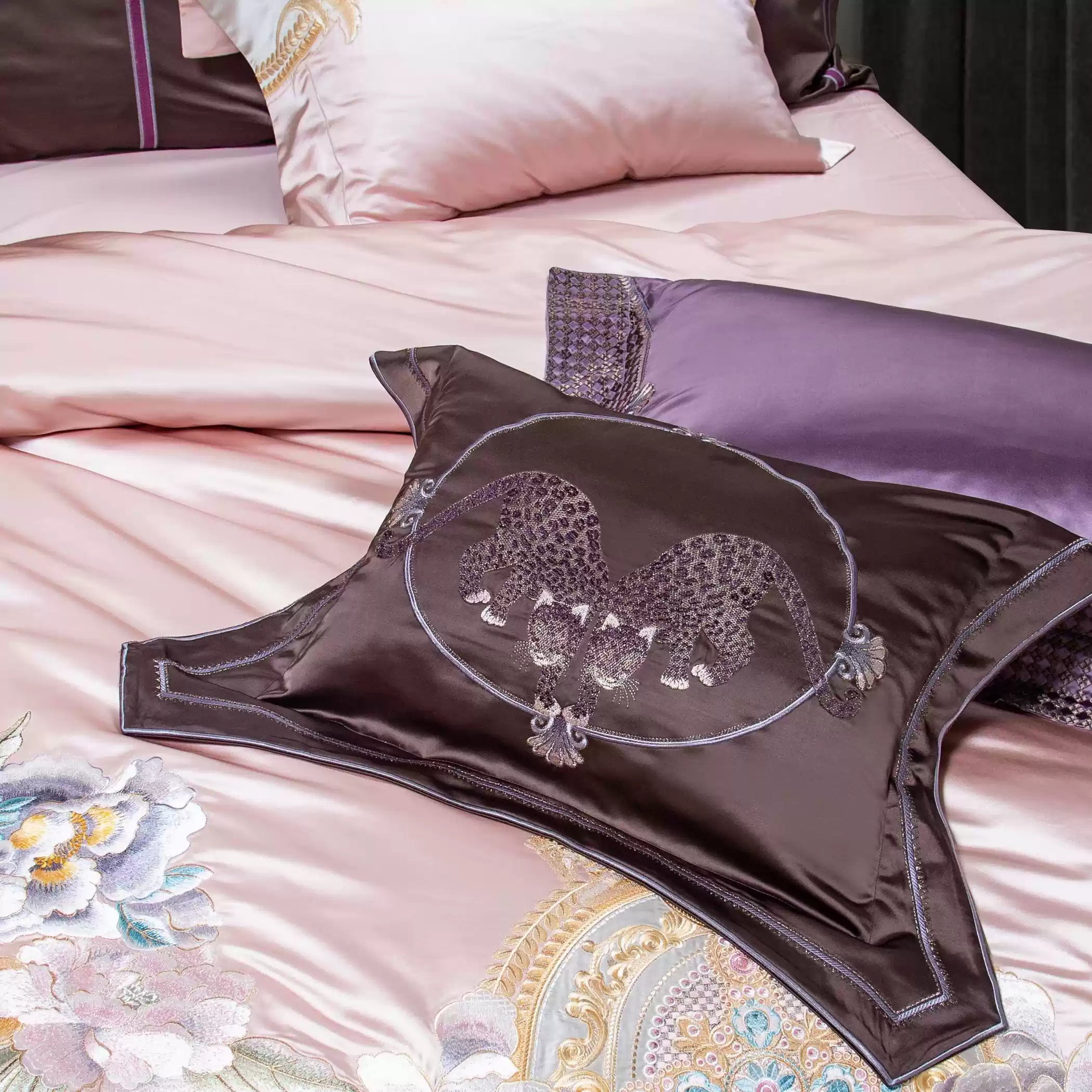 A detailed view of a luxury bedding set featuring a dark brown decorative pillow with an embroidered twin leopard design framed in a circular pattern. The backdrop includes soft pink and lavender silk pillows, accented with floral embroidery and gold embellishments, enhancing the set's opulence and intricate artistry.