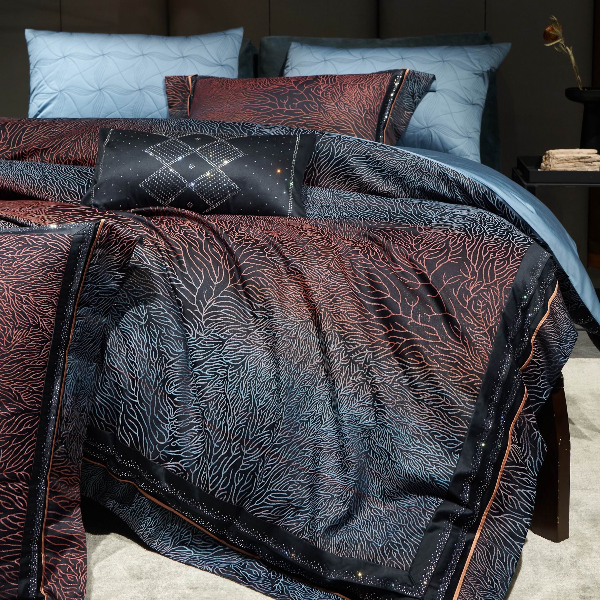 A detailed view of a luxury bedding set featuring a coral-inspired pattern in black, navy, and red tones. The intricate design flows elegantly across the duvet cover and pillow shams, with a gradient effect transitioning between dark and lighter hues. The bedding is paired with a black decorative pillow adorned with sparkling geometric embroidery, adding a sophisticated and contemporary touch. Soft blue sheets and pillows in the background 