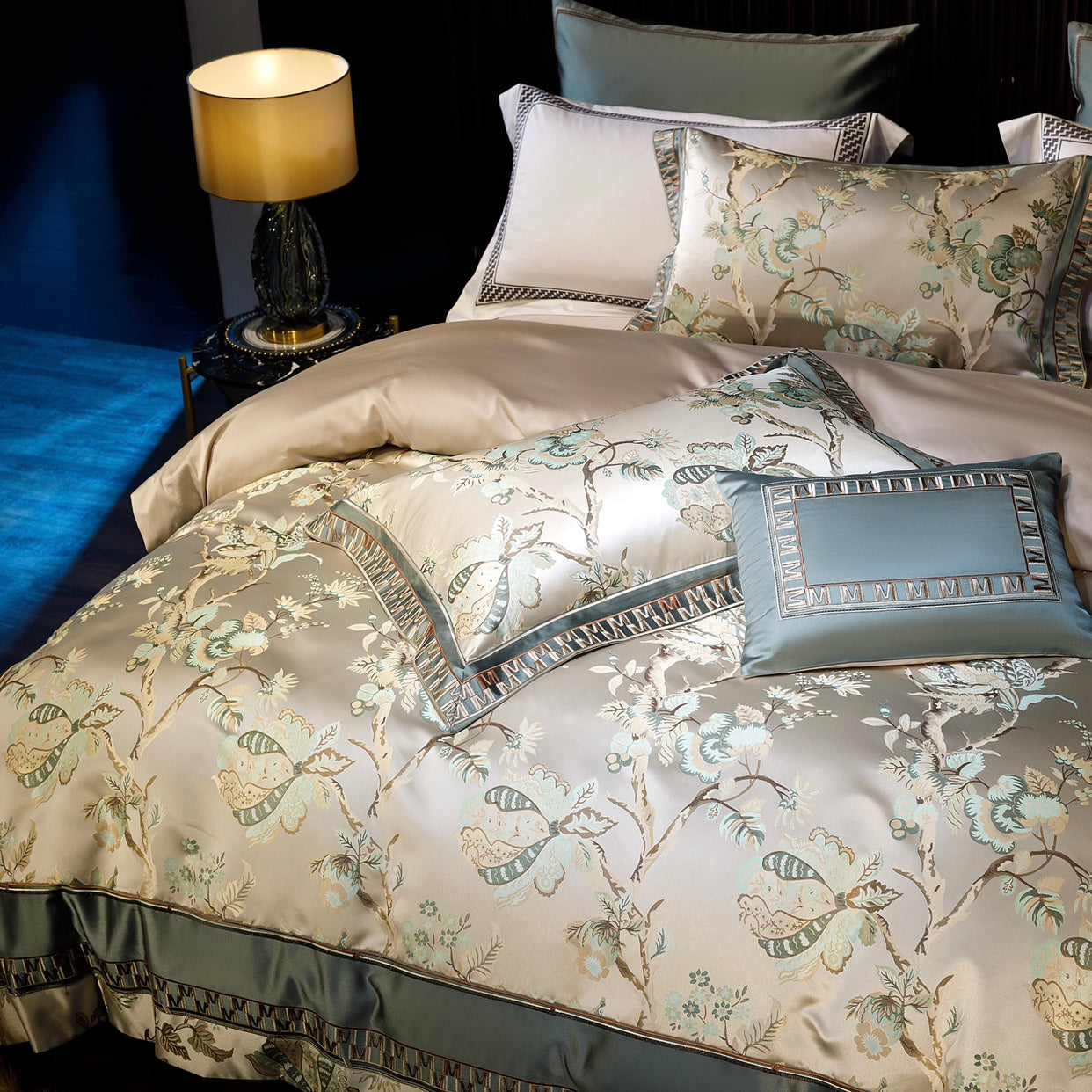 This close-up image highlights an elegant bedding set adorned with intricate floral and butterfly motifs in a soothing beige and sage green color palette. The bedding exudes luxury with its silky finish and detailed embroidery, complemented by geometric accents along the edges. A matching rectangular throw pillow with a framed border design adds a sophisticated touch. Soft beige sheets provide a harmonious backdrop, while a stylish black and gold side table holding a lamp with a warm glow enhances the overa