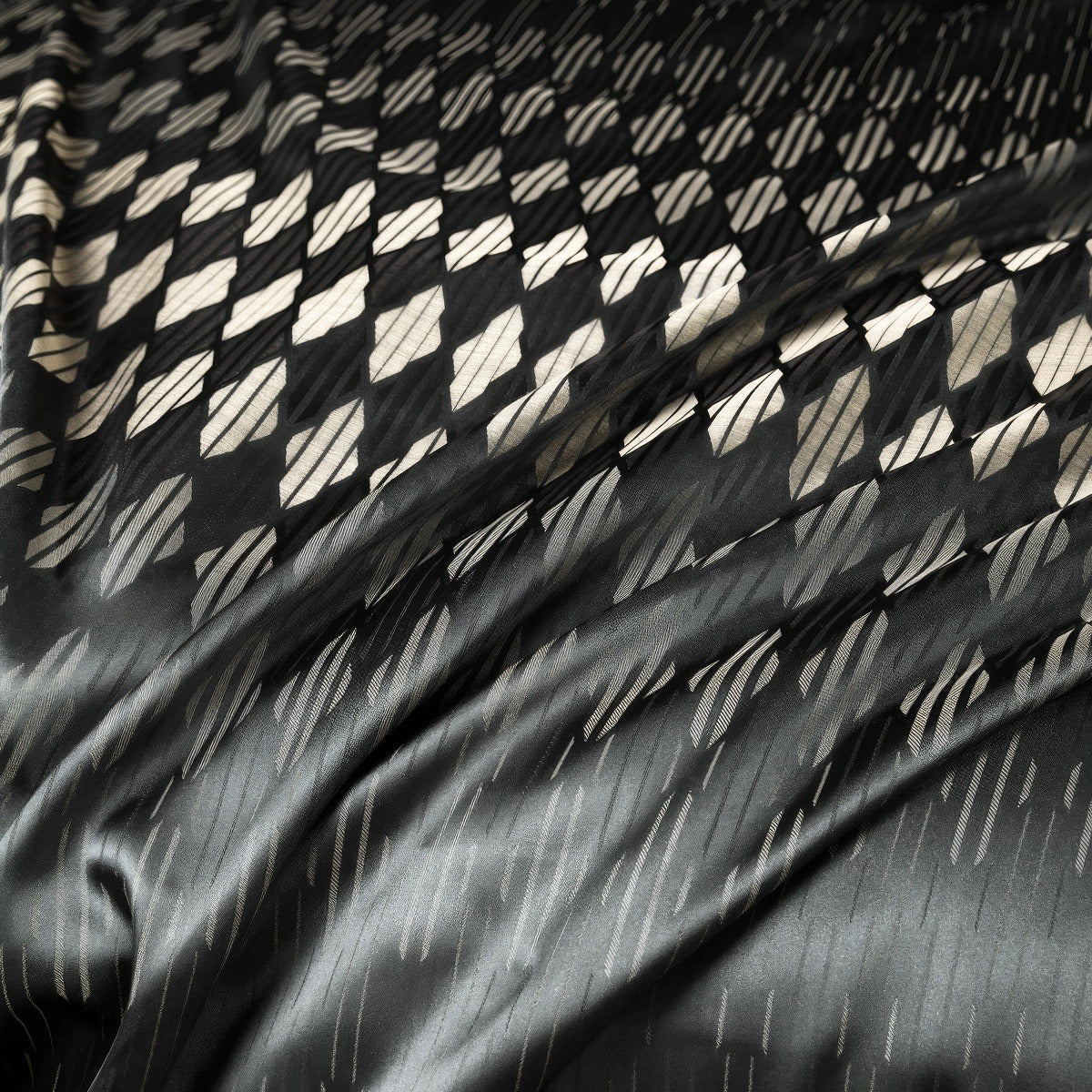 Close-up of luxurious black fabric with an intricate geometric diamond pattern in contrasting beige and metallic tones, creating a shimmering effect.

