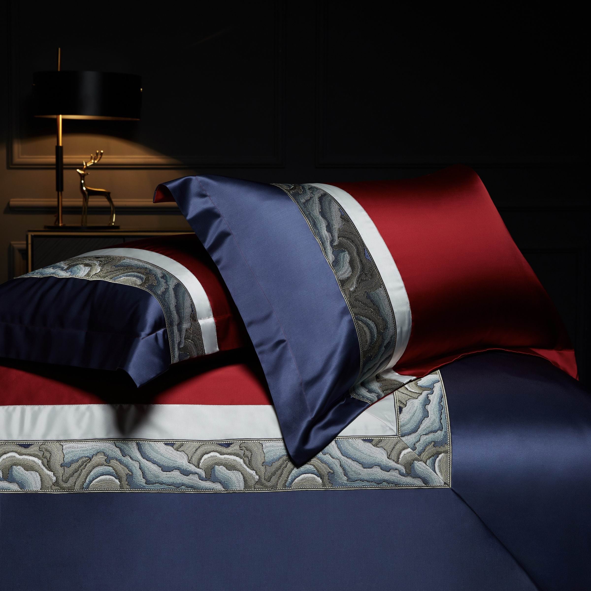 Two pillowcases with deep red and navy blue colors, featuring wave-like embroidery on a white trim, placed on a bed against a dark background with a gold lamp and decor piece.