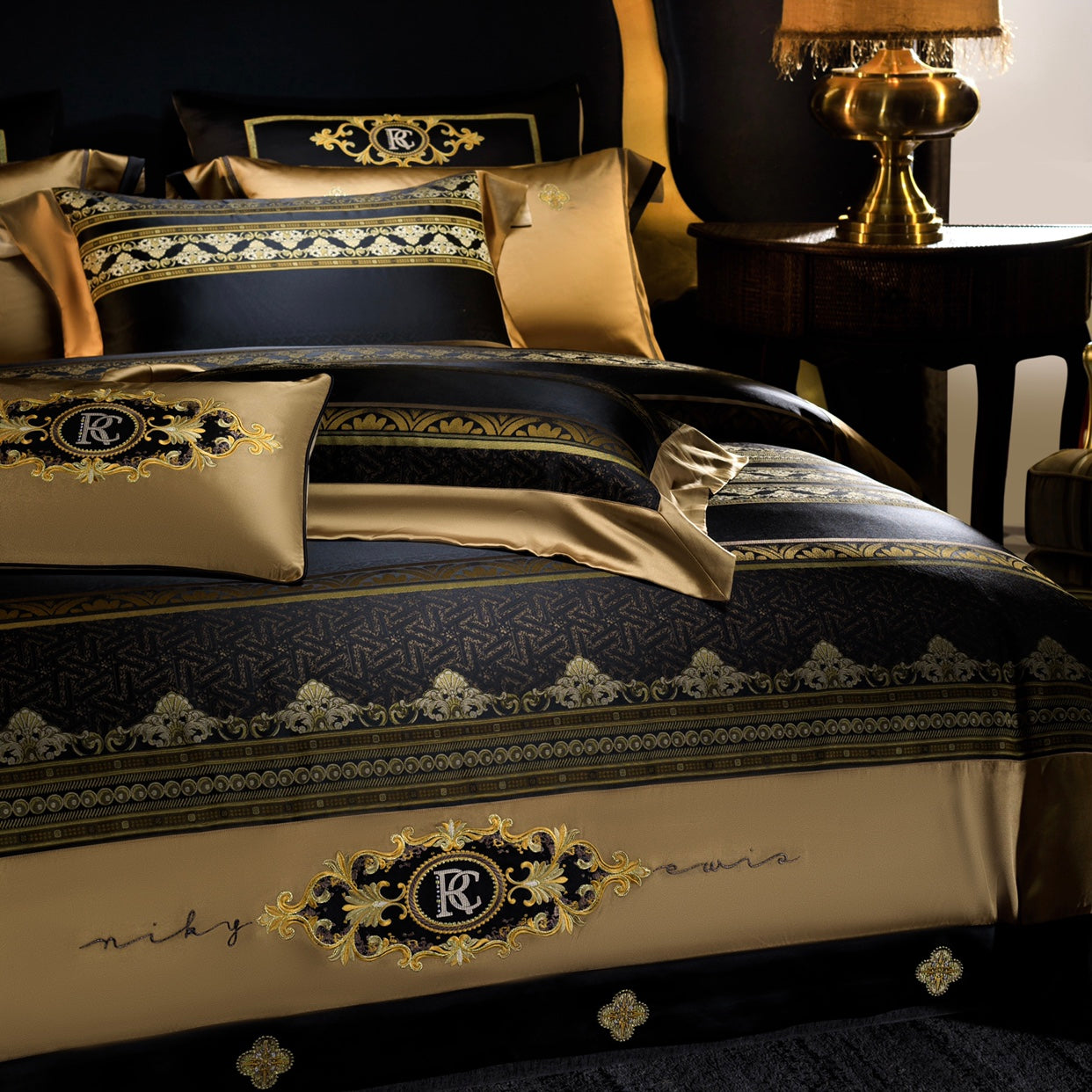 A close-up view of a luxurious black and gold bedding set featuring intricate baroque embroidery and regal designs. The bedding includes a monogrammed crest as a focal point, bordered by ornate golden patterns on the duvet cover and pillowcases. A decorative cushion with matching embroidery enhances the elegant aesthetic. In the background, a gold-toned table lamp on a dark wooden side table adds warmth to the sophisticated ambiance, while the rich textures of the bedding exude opulence and refinement.