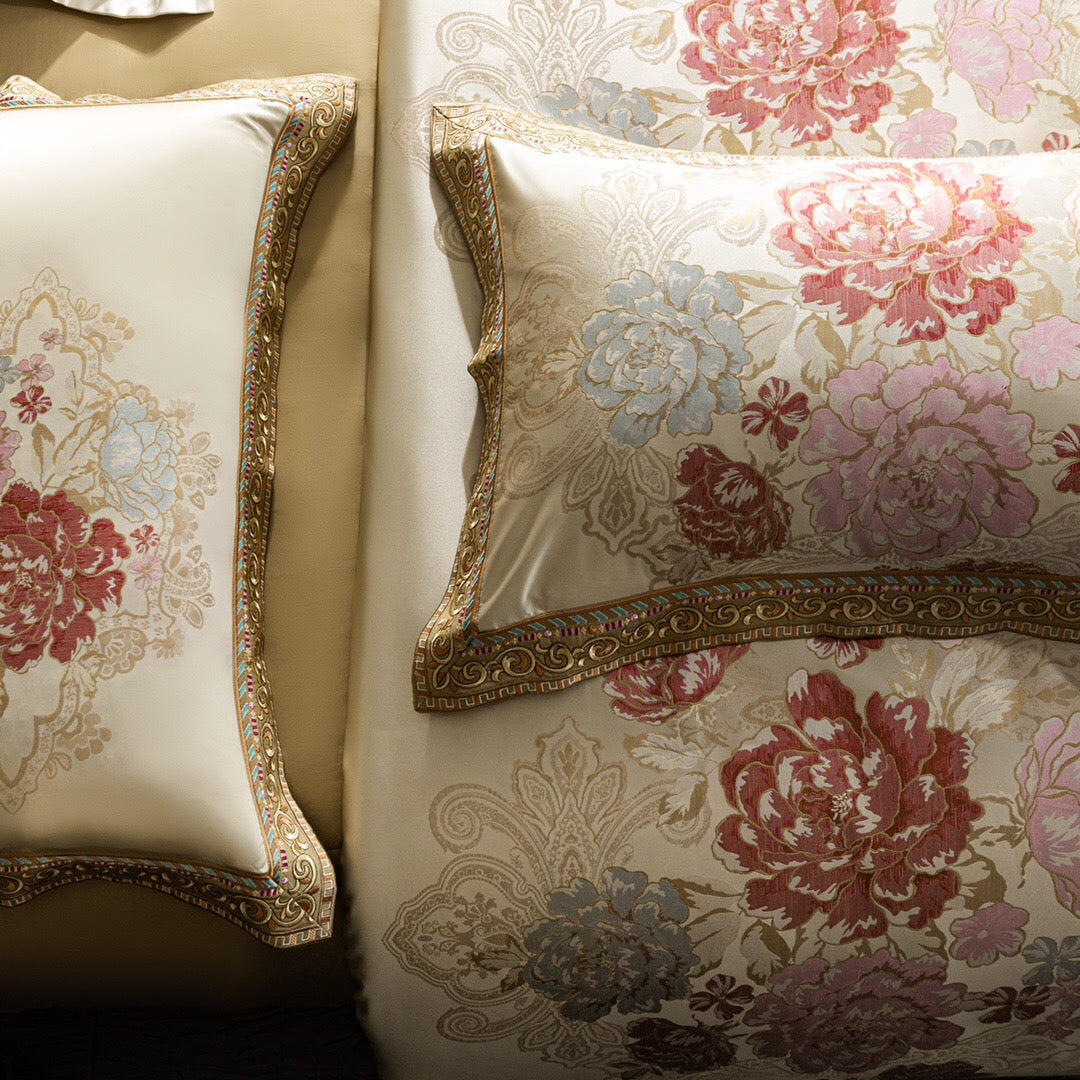 A close-up view of a luxurious ivory bedding set showcasing pillowcases and a duvet cover adorned with exquisite floral embroidery. The embroidery features intricate patterns of peonies in shades of pink, red, and light blue, framed by gold-accented borders with ornate details. The delicate embroidery highlights the craftsmanship and opulence of the design, making this bedding set a statement of elegance and sophistication.