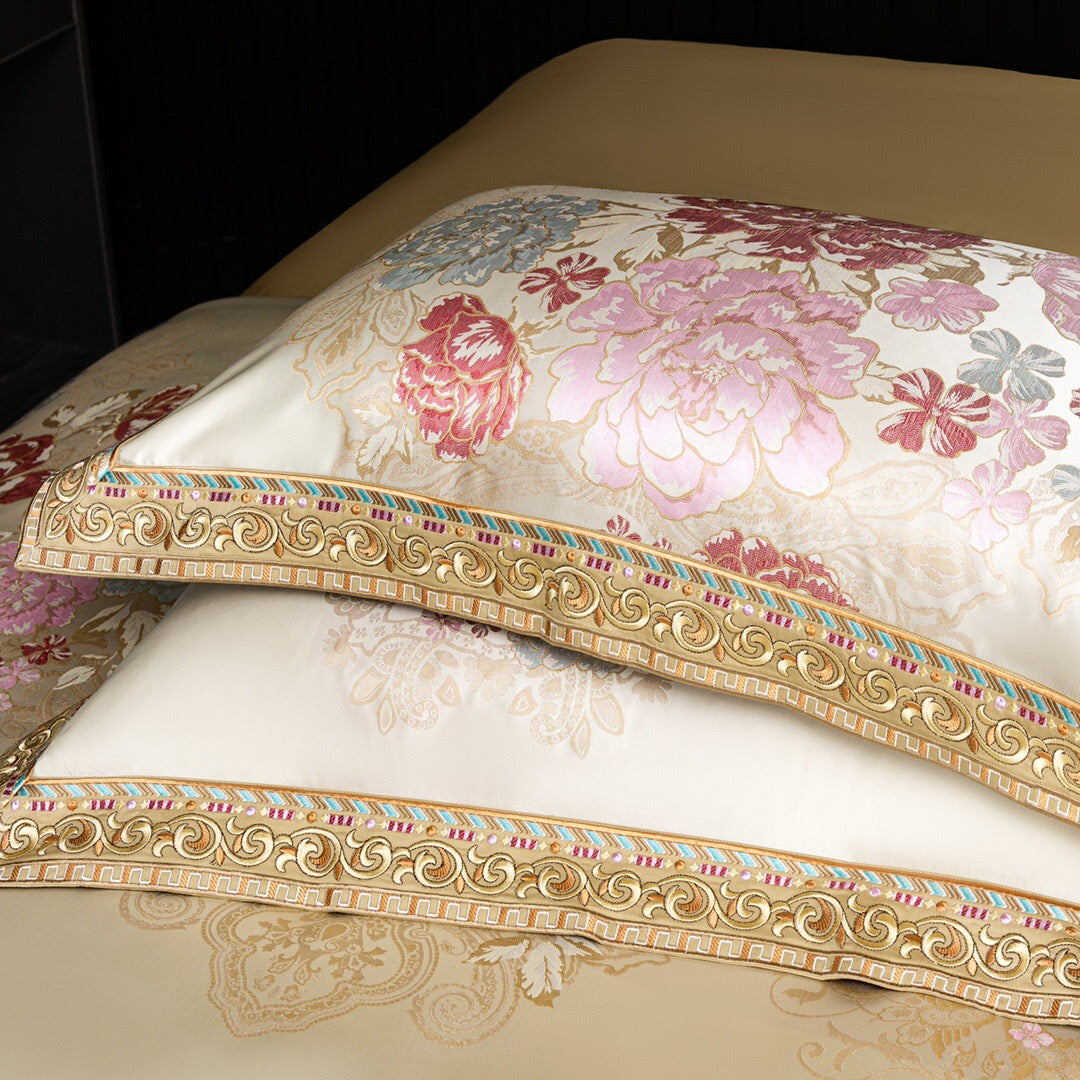 A detailed close-up of ivory pillowcases featuring luxurious floral embroidery in shades of pink, red, and light blue. The intricate designs are framed with an ornate golden border, accented by colorful detailing that enhances the richness of the pattern. The craftsmanship and opulence of these pillowcases add an elegant touch, making them a standout feature in a premium bedding set.