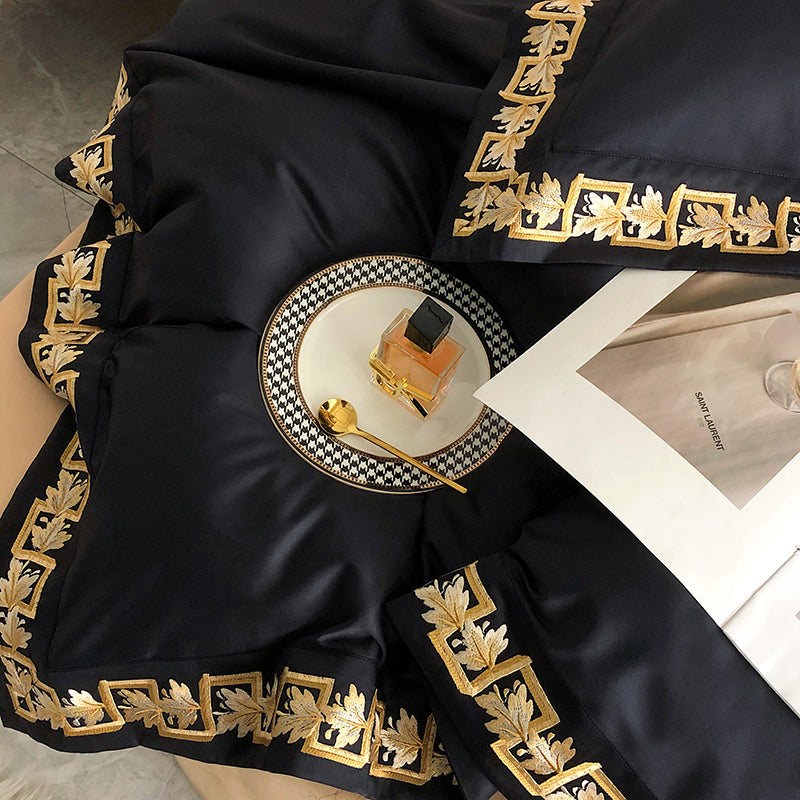 Detailed view of black Egyptian cotton fabric with intricate gold embroidery, complemented by a golden spoon, perfume bottle, and designer plate, showcasing a luxurious and refined aesthetic.

