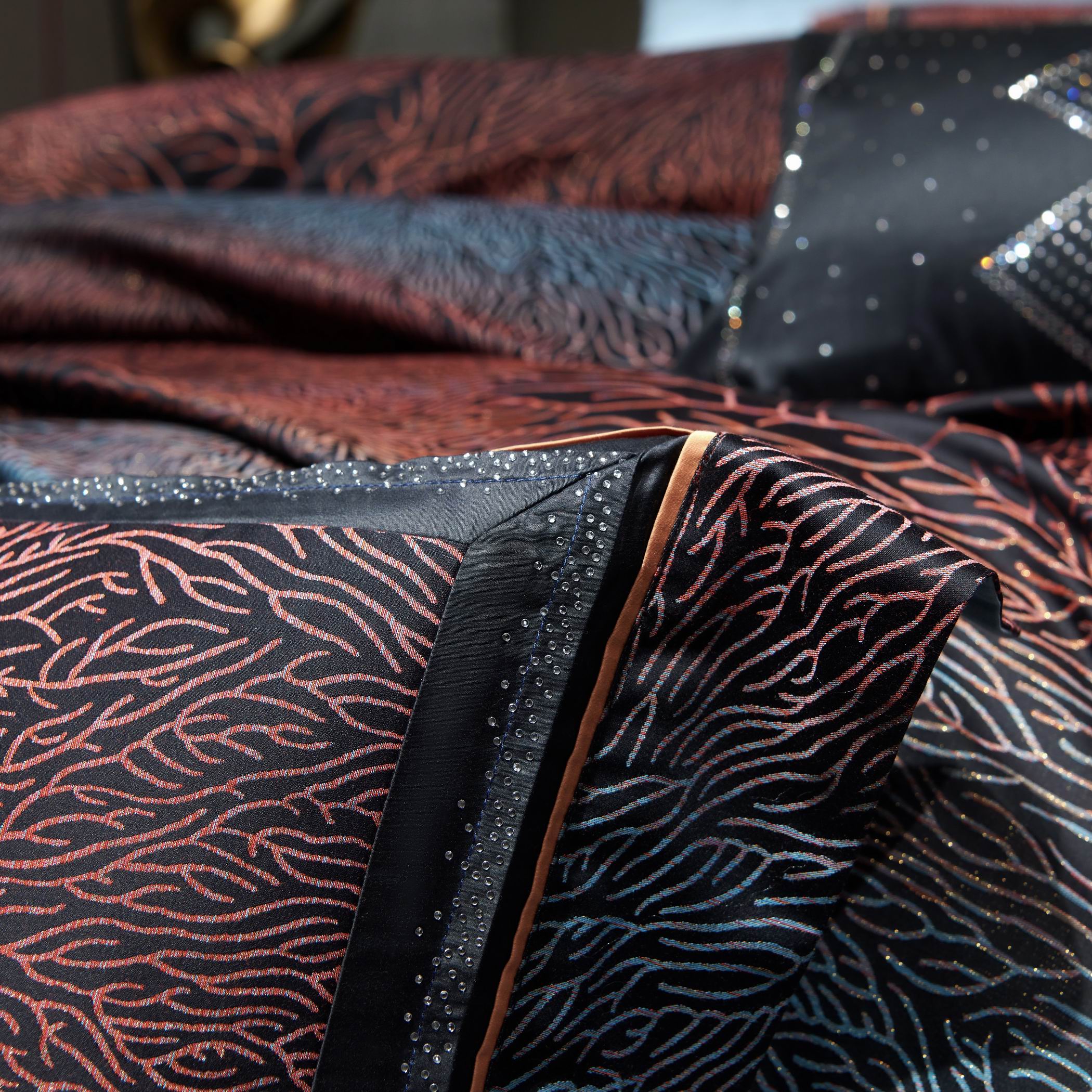 A close-up view of a luxury bedding set featuring a detailed coral-inspired pattern in shades of red, orange, and blue against a black base. The intricate design is accentuated by shimmering embellishments, adding a touch of sparkle. A fine satin border with contrasting piping enhances the edges, creating a sophisticated and polished look. The texture and luster of the fabric further emphasize the high-end craftsmanship, ideal for an opulent and stylish bedroom setting.

