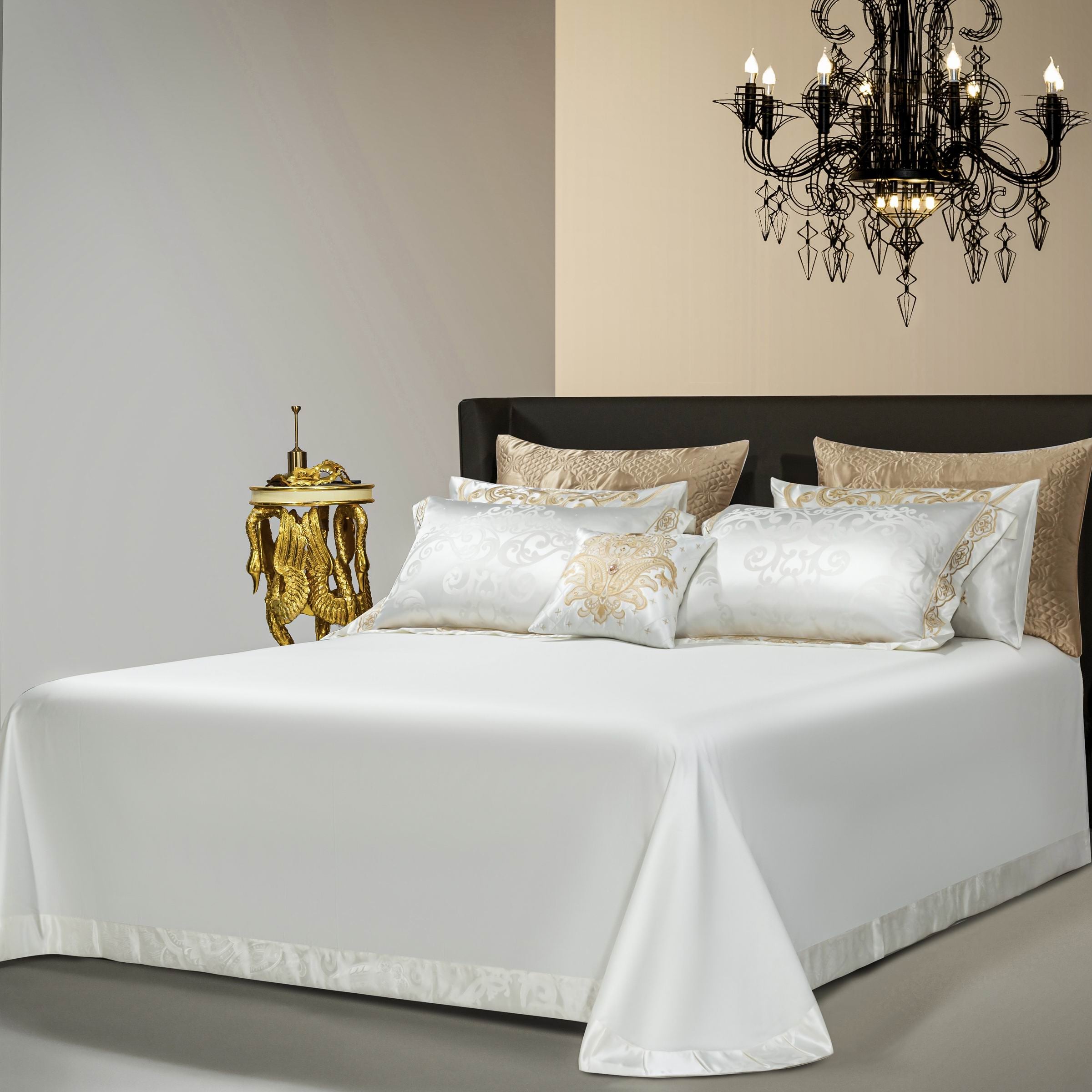 A refined bedroom setup showcasing a white jacquard bedding ensemble with intricate gold embroidery on the pillowcases and duvet edges. The bedding exudes luxury, complemented by textured golden accent pillows at the back. A decorative chandelier above adds a touch of grandeur, while a gold ornamental side table beside the bed enhances the opulent ambiance of the room.

