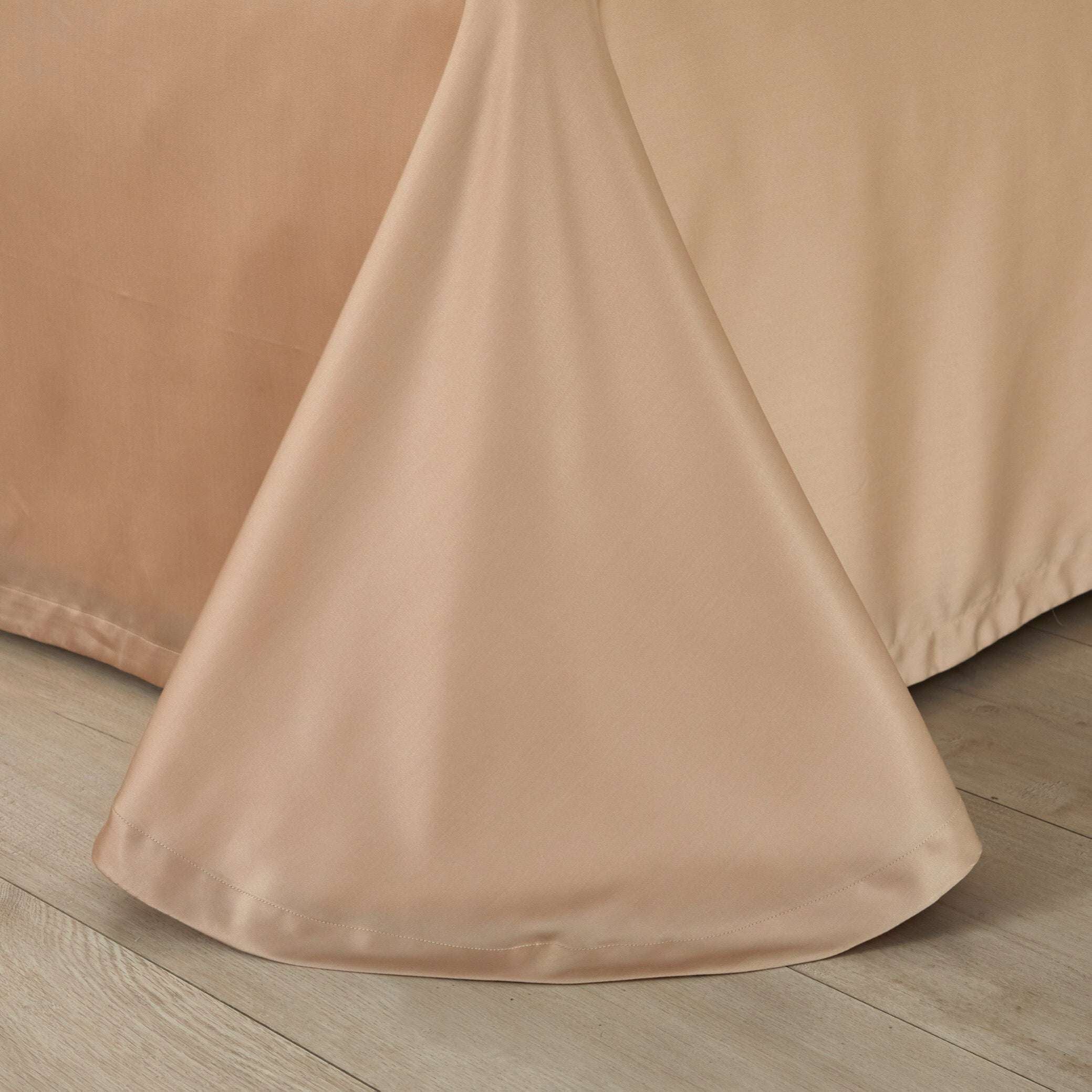 A close-up of a champagne gold silk bed skirt gracefully flowing to the floor. The luxurious fabric highlights a smooth, seamless texture with a subtle sheen, adding sophistication to the bedding ensemble. The hemline is neatly stitched, ensuring durability and a polished finish.