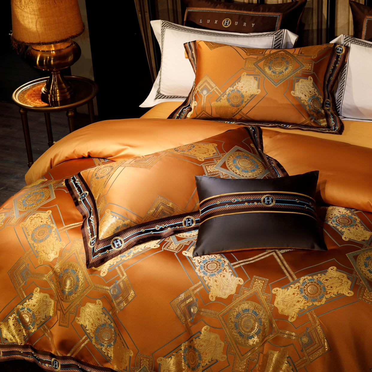 A vibrant orange bedding set adorned with elaborate gold and blue geometric motifs, complemented by dark brown borders featuring embroidered "H" monograms in blue and gold. The glossy fabric enhances the luxurious design, paired with accent pillows showcasing coordinated patterns and elegant detailing. The setup exudes a regal and sophisticated ambiance, perfect for an opulent bedroom aesthetic.