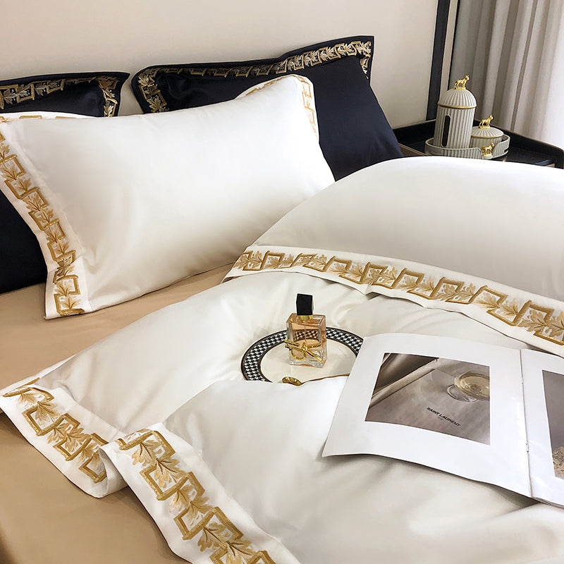 Luxurious white Egyptian cotton bedding accented with intricate gold embroidery, paired with contrasting black pillows, a designer perfume bottle on a houndstooth-patterned plate, and an open luxury magazine.

