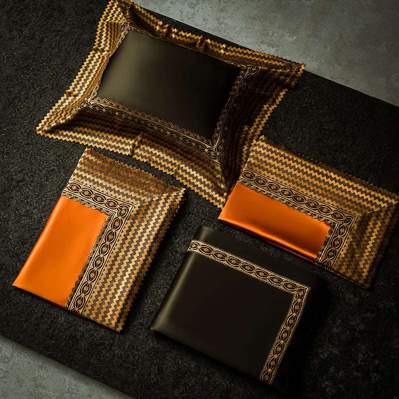 Two sets of luxurious pillowcases and bedding sheets laid flat, featuring contrasting orange and dark green satin with intricate gold and black embroidered borders. The pieces are artfully arranged on a dark textured surface, highlighting the bold, opulent design.

