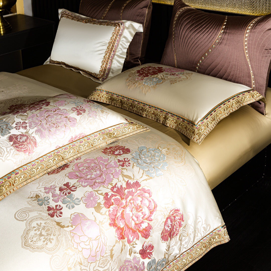 An elegant close-up of a luxurious bedding set featuring an ivory duvet cover with intricate floral patterns in pastel pink, blue, and red tones. The edges are adorned with ornate golden embroidery and colorful accents. Coordinating pillows, including a quilted mauve design and others with matching floral motifs and gold trim, rest on a beige bedspread. The composition highlights the fine detailing and opulence of the fabric, creating a sophisticated and regal bedroom ambiance.