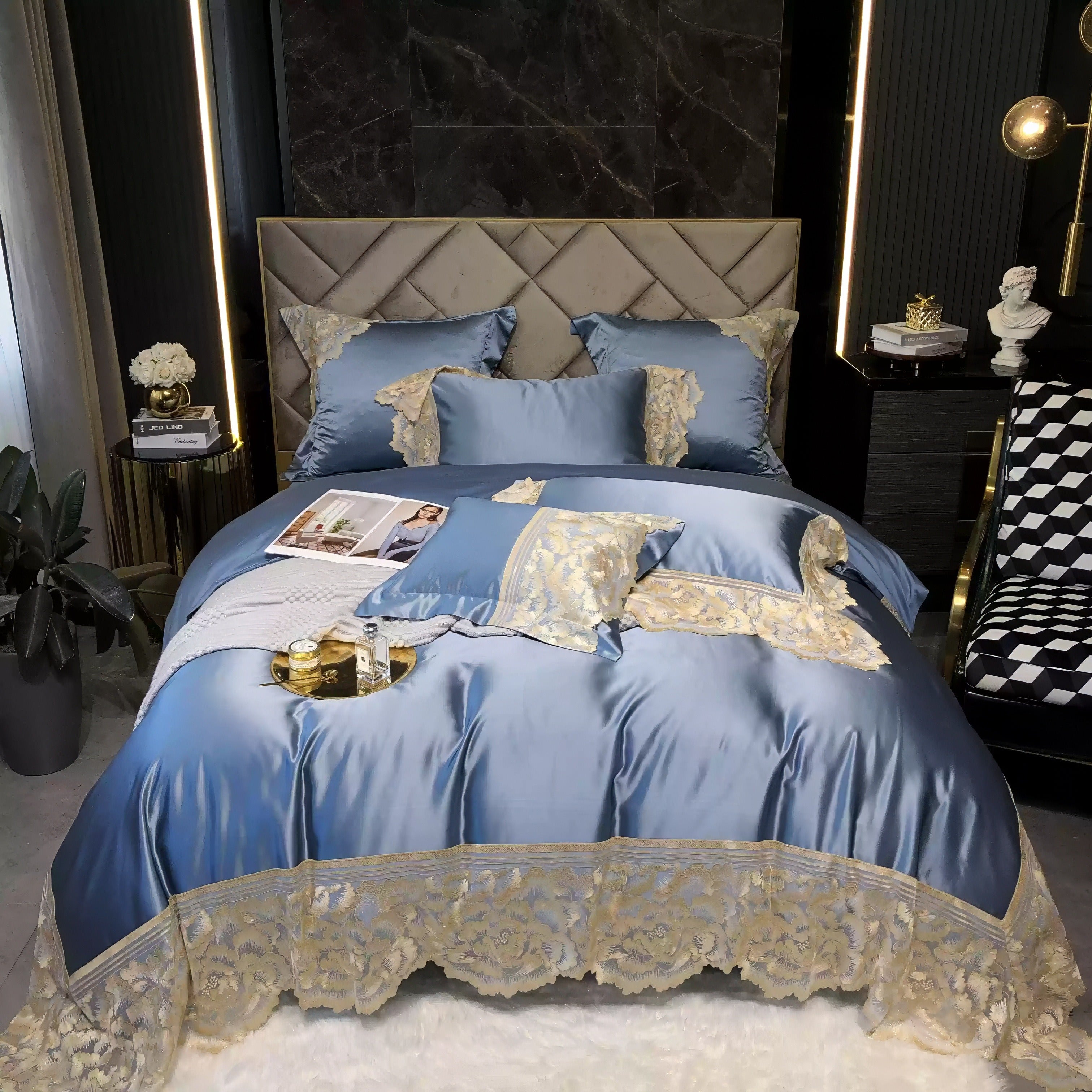 Luxurious blue satin bedding set with intricate gold lace trim, matching pillows, and decorative accents on a tufted headboard bed, complemented by a cozy throw and magazine.

