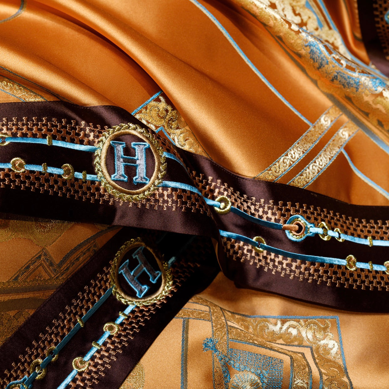 A close-up of a luxurious orange fabric with intricate golden patterns and contrasting brown borders adorned with blue and gold embroidery. The border features a monogrammed "H" encased in a circular design, accented by chain-like details in blue and gold. The vibrant and rich details highlight the opulent craftsmanship of the bedding.