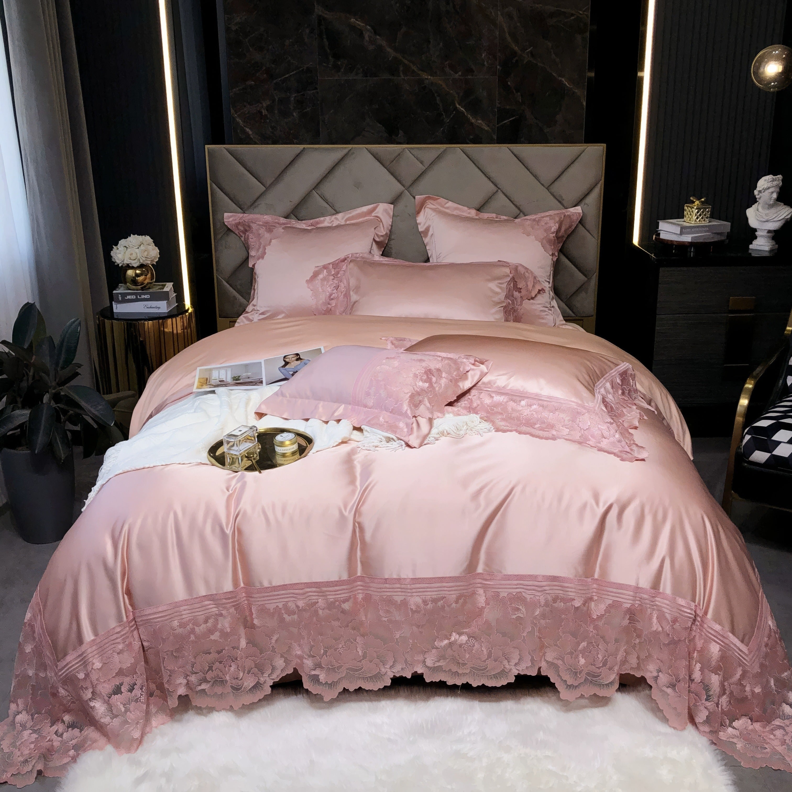 Pink satin bedding set with intricate lace detailing along the edges, featuring matching pillows and decorative cushions on a plush bed in a modern, luxurious bedroom.

