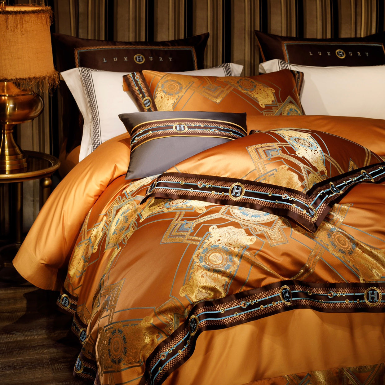 A luxurious bedding set in vibrant orange with intricate golden patterns and blue highlights, framed by a bold brown border featuring monogram details. The pillows and duvet showcase elegant geometric designs, paired with white pillowcases featuring black decorative trim. The setup is enhanced by a golden table lamp with a fringed shade and a striped upholstered headboard, creating a sophisticated and opulent ambiance.