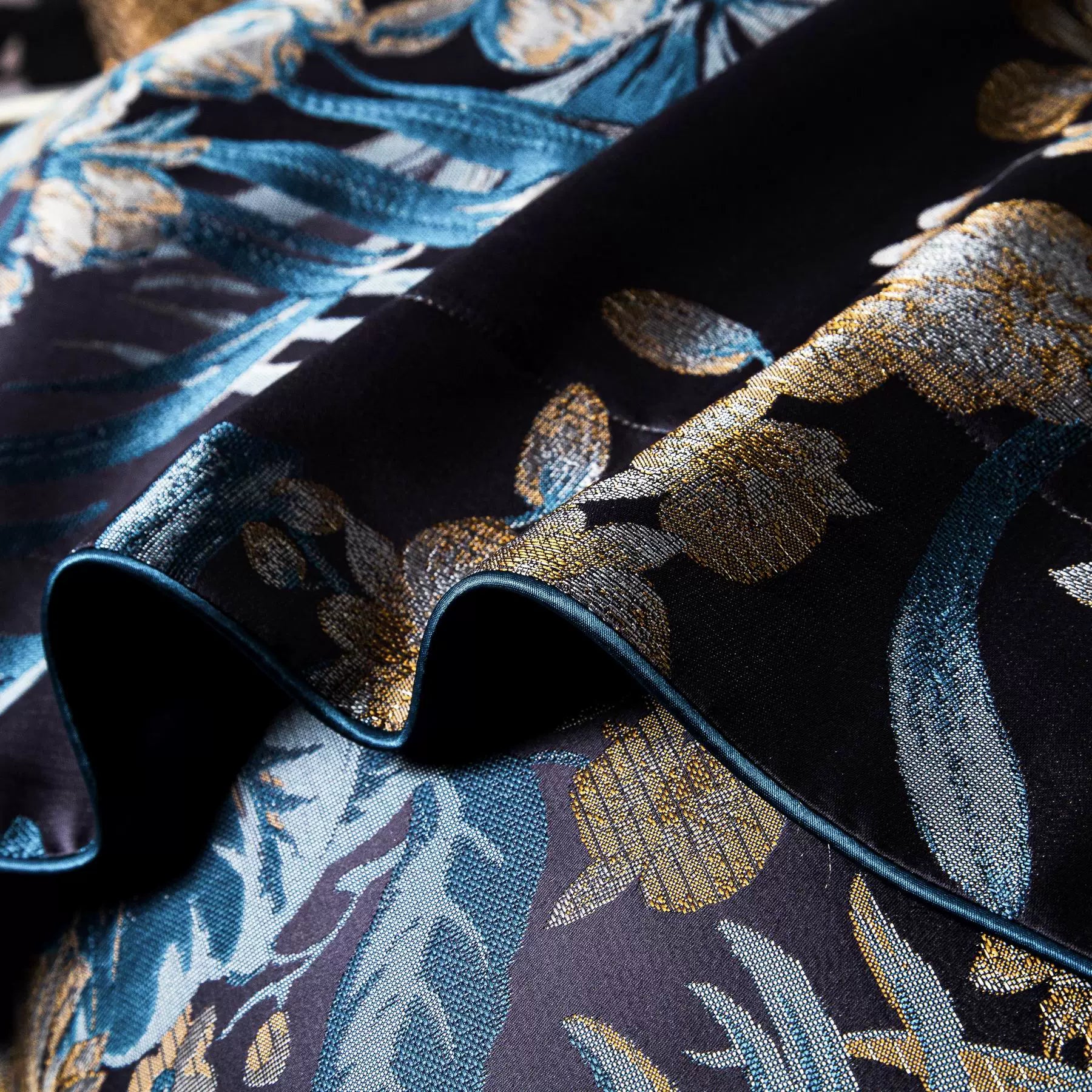 This image showcases a close-up of the pillowcase from the luxurious bedding set. The intricate floral patterns in gold and teal hues stand out beautifully against the deep navy background. The design highlights the rich texture and fine detailing, with the piping adding a sophisticated finishing touch. This bedding exudes elegance and is ideal for a refined and stylish bedroom look.