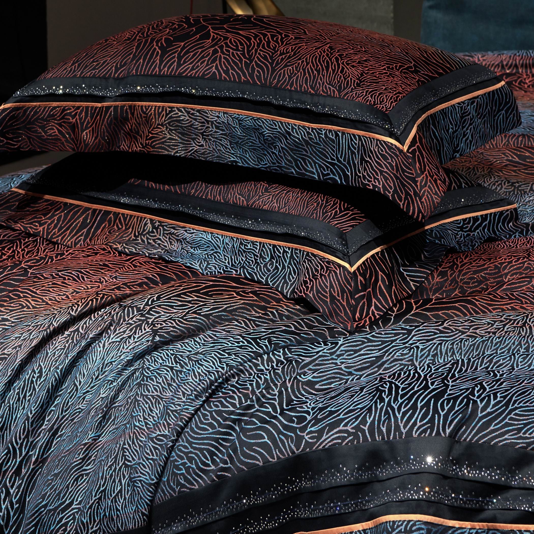 A luxurious close-up of a bedding set with intricate coral-inspired embroidery in shades of red, orange, and blue against a sleek black background. The pillowcases are adorned with shimmering accents along the edges, framed by a delicate piping detail for a refined look. The elaborate stitching and color transitions flow seamlessly across the fabric, enhancing its opulent, textured appearance. This high-end bedding set showcases meticulous craftsmanship and a contemporary design, ideal