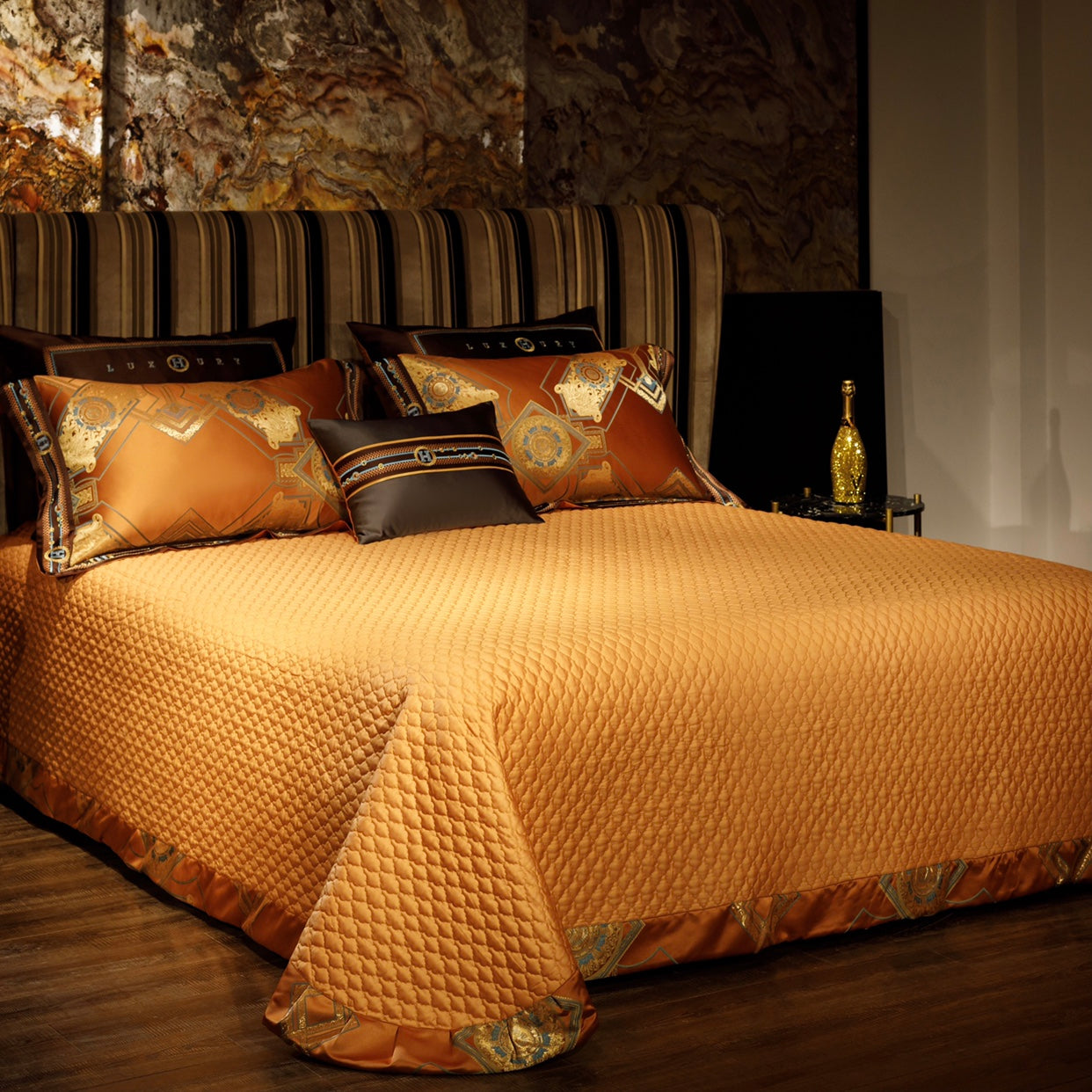 A quilted orange bedspread with a textured pattern covering the bed, complemented by matching pillowcases adorned with intricate gold and blue geometric designs and dark brown accents. The setup includes a striped upholstered headboard and gold-tone decor elements, creating a luxurious and warm bedroom aesthetic with rich tones and elegant details.