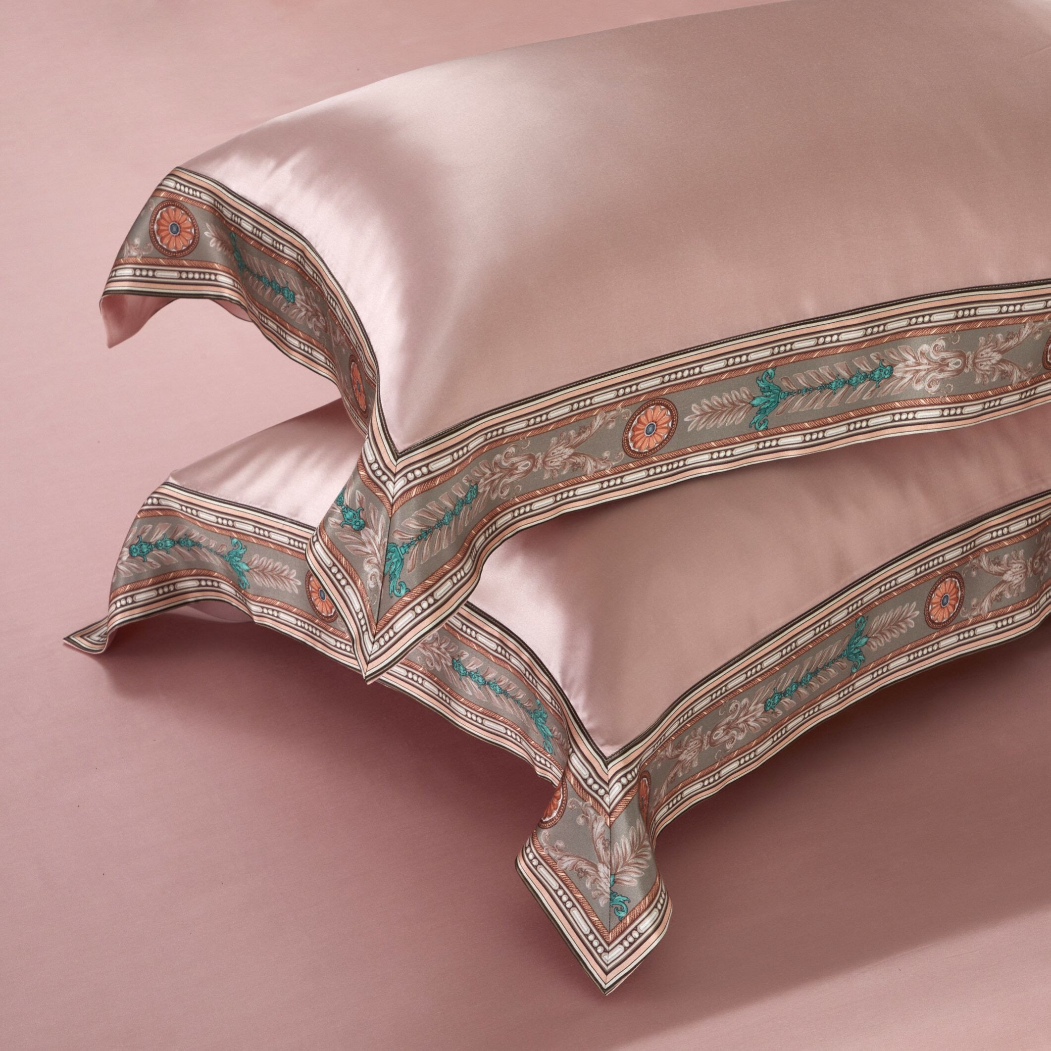 Two elegant blush pink pillowcases embellished with detailed ornamental borders featuring intricate floral and geometric patterns in soft earth tones and teal accents. The luxurious fabric gleams with a smooth finish, set against a matching pink backdrop, exuding sophistication and opulence.