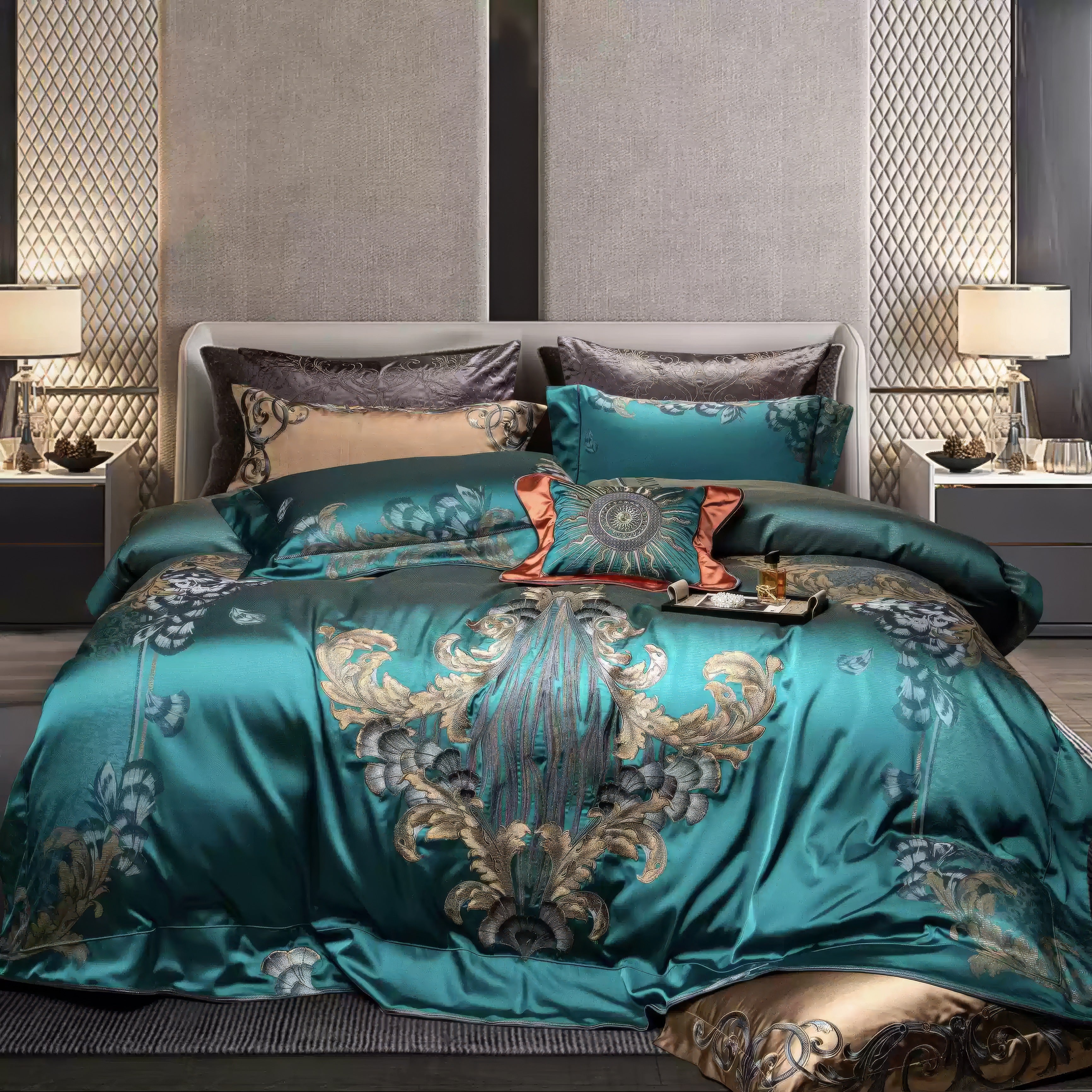 Luxurious bedding in a teal satin finish adorned with intricate gold and silver embroidery, featuring floral and baroque motifs. A decorative orange and teal throw pillow with embroidered detailing serves as a centerpiece on the bed. The headboard is upholstered in light gray, flanked by modern bedside tables with table lamps and minimalistic decor, including metallic accents and glassware. The quilted wall panels add depth and sophistication to the room.