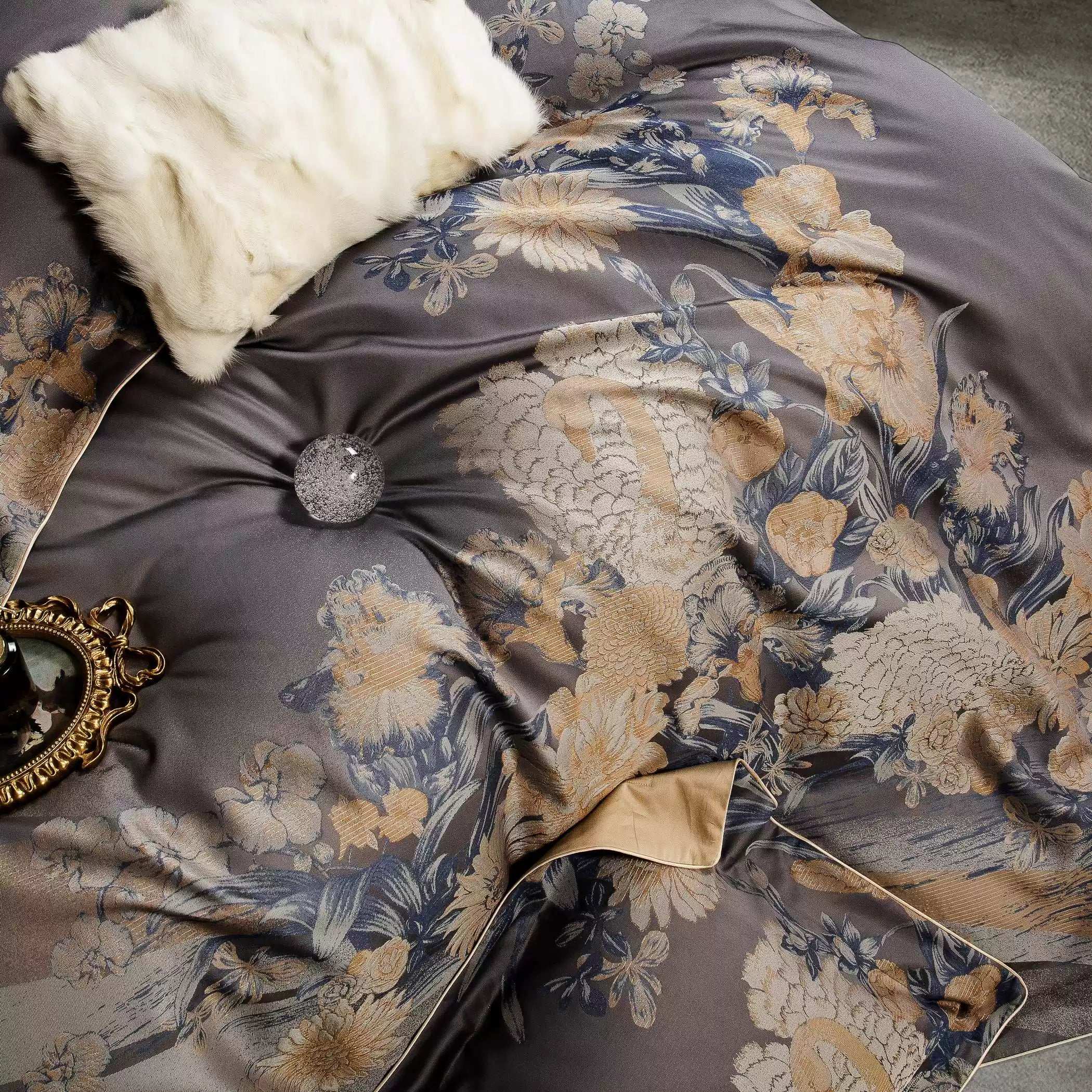 A detailed view of a dark gray bedding set adorned with intricate gold and blue floral patterns. The design highlights a tufted center with a sparkling glass accent, creating a luxurious texture. A fluffy white decorative pillow and a gold-framed mirror enhance the overall elegance, while a contrasting gold-tone lining peeks from the folded corner of the duvet.