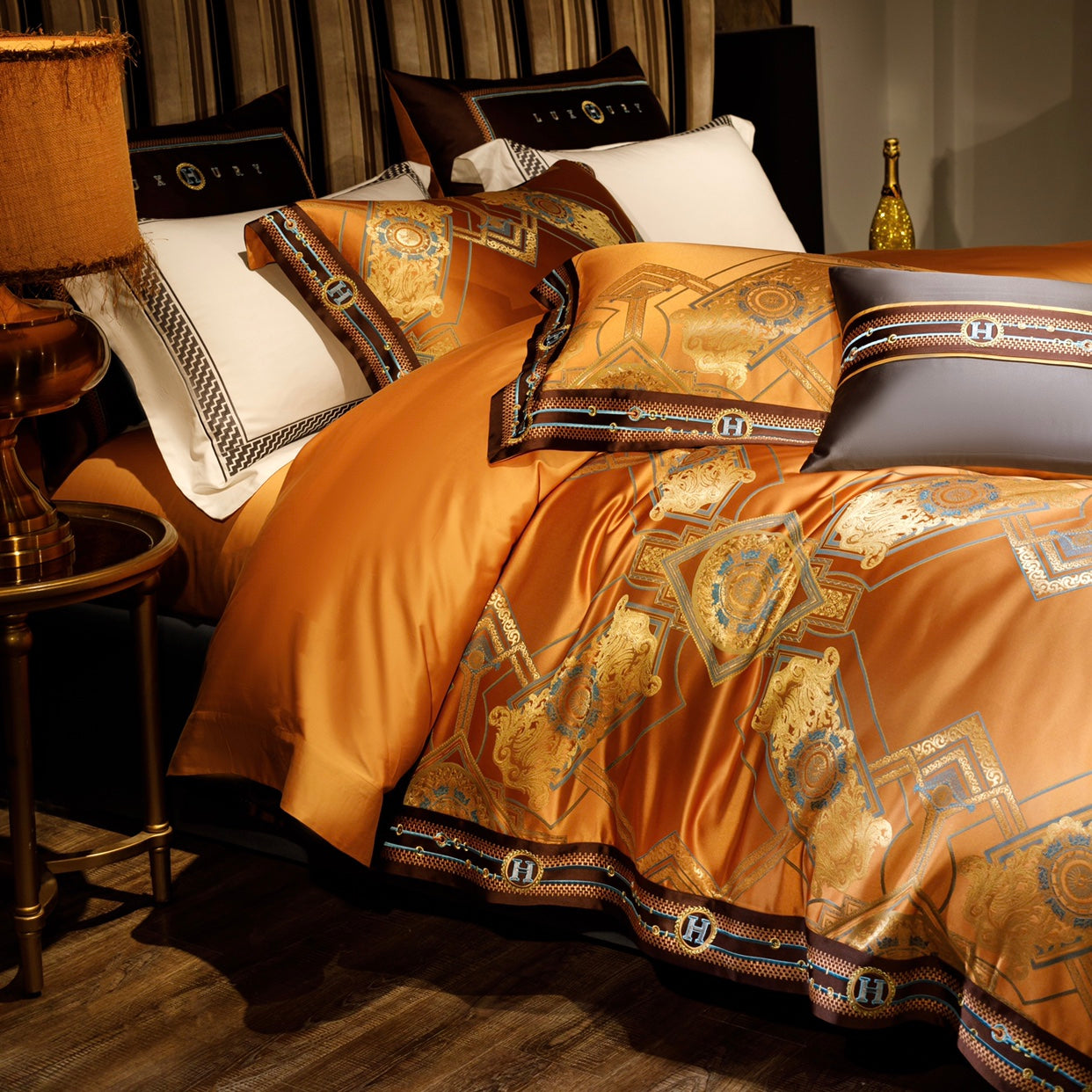 An elegant close-up of a luxury bedding set in warm orange hues, highlighted with golden geometric and intricate blue accents. The design features ornate patterns bordered with contrasting elements in brown and black, complete with monogram details. The setup is complemented by a white pillowcase with a black geometric trim, a golden decorative bottle, and a classic lamp on a wooden side table, creating a regal and sophisticated atmosphere.