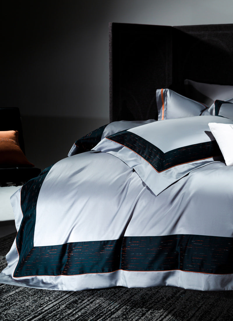 Close-up of a luxurious silver duvet cover with contrasting deep navy blue borders accented by a subtle red stitch pattern. The rich fabric drapes elegantly over the bed, showcasing its smooth texture and high-end design. A portion of matching pillowcases with the same navy and red border detail is visible against the headboard. Soft lighting highlights the duvet’s sheen and the intricate stitching, enhancing the sophisticated, modern aesthetic of the bedroom.

