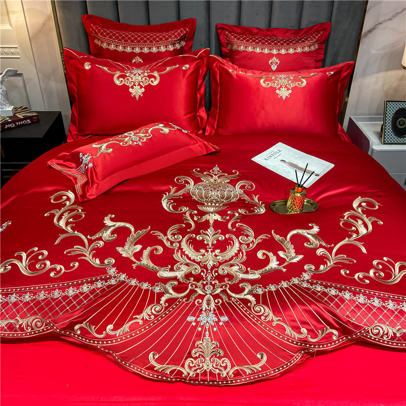 Close-up of a red Egyptian cotton bedding set with intricate gold embroidery. The design features ornate floral and scrollwork patterns, highlighted with shimmering gold threads across the duvet and matching pillowcases. The vibrant red fabric showcases a silky sheen, emphasizing the luxurious quality of the Egyptian cotton material. The bedding is accessorized with a gold tray holding a reed diffuser and a magazine for a sophisticated and elegant presentation.

