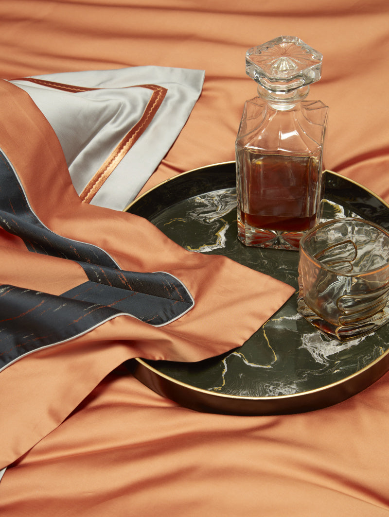 A luxurious bedding setup featuring silky orange fabric with contrasting black and gold trim. A dark marble tray holds a glass decanter filled with amber liquid and a matching tumbler, complementing the opulent aesthetic. A white pillow with delicate orange piping peeks through the folds, adding refinement to the scene.

