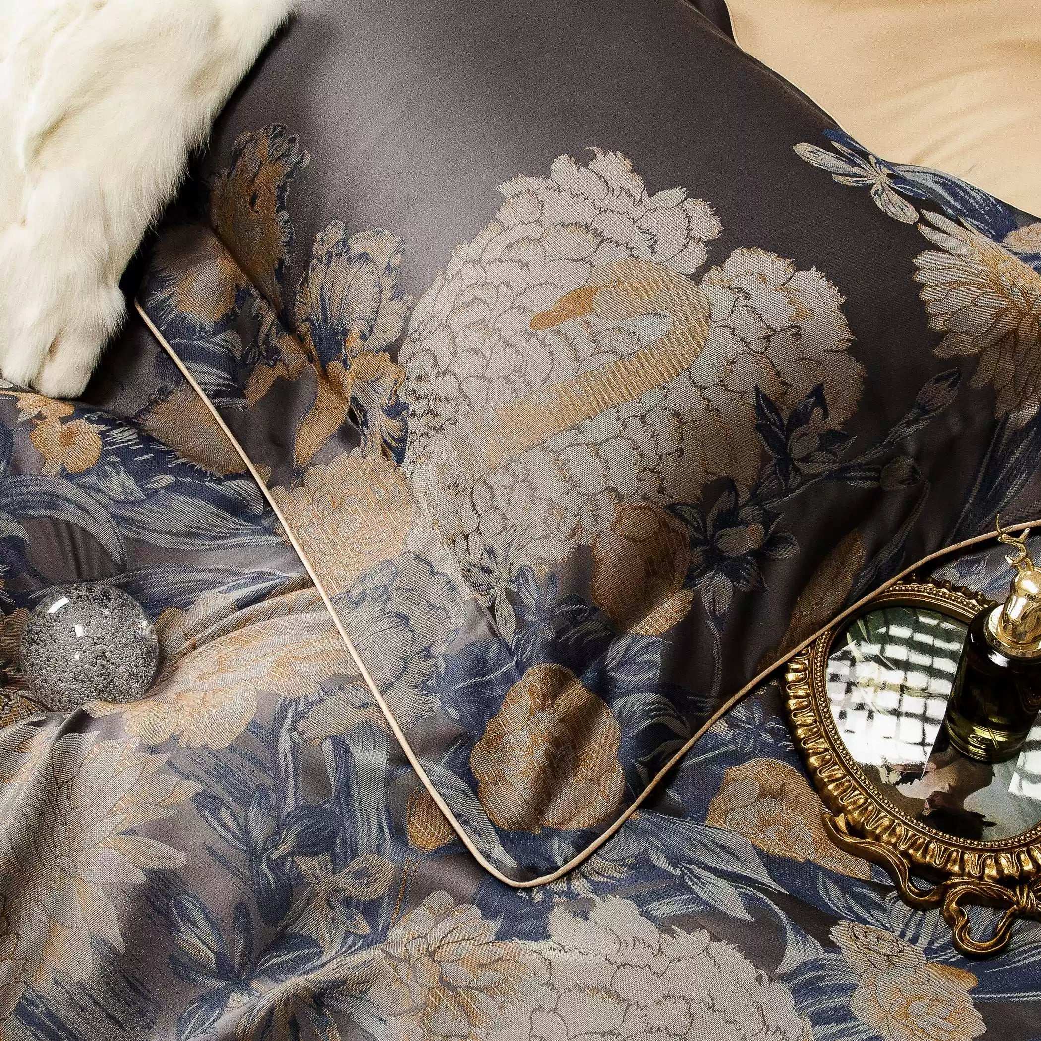 Close-up of a dark gray bedding set featuring an intricate embroidered swan amidst detailed gold and blue floral designs. The fabric's sheen enhances the richness of the embroidery, while a fine gold trim outlines the edges. A white faux fur pillow adds texture, and a gold-framed mirror with a perfume bottle reflects sophistication. A subtle glittering accent completes the elegant composition.