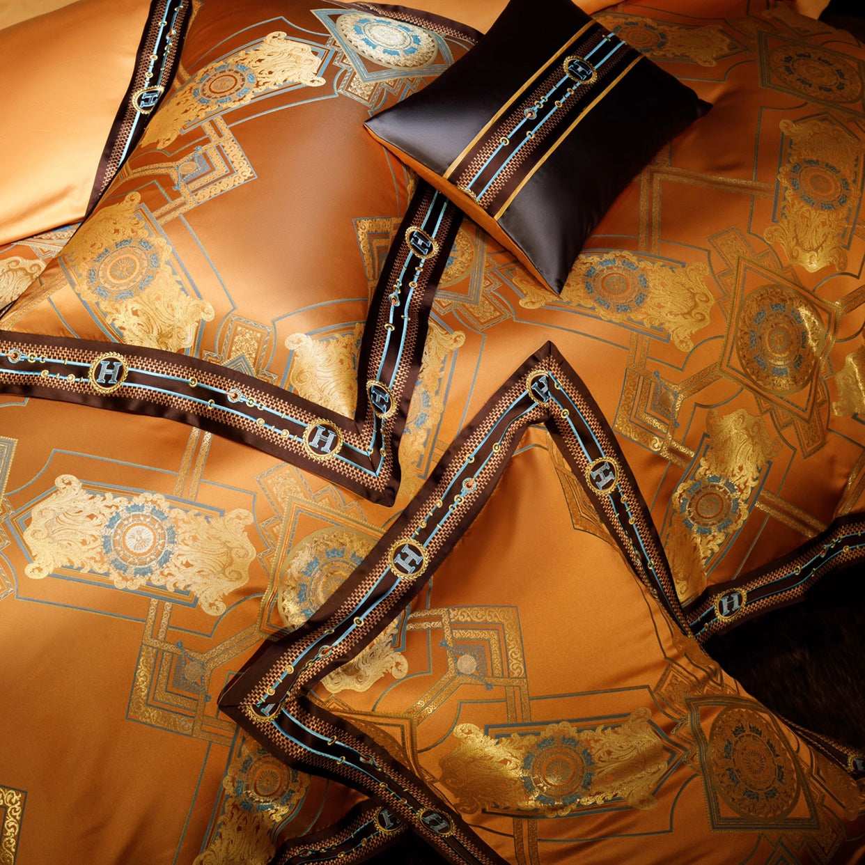 A luxurious bedding set in a rich orange hue with intricate golden patterns and ornate designs. The design is accented with dark brown borders featuring monogrammed "H" motifs in blue and gold embroidery, creating a regal and elegant appearance. The fabric displays a glossy finish, emphasizing its premium quality and sophisticated craftsmanship.