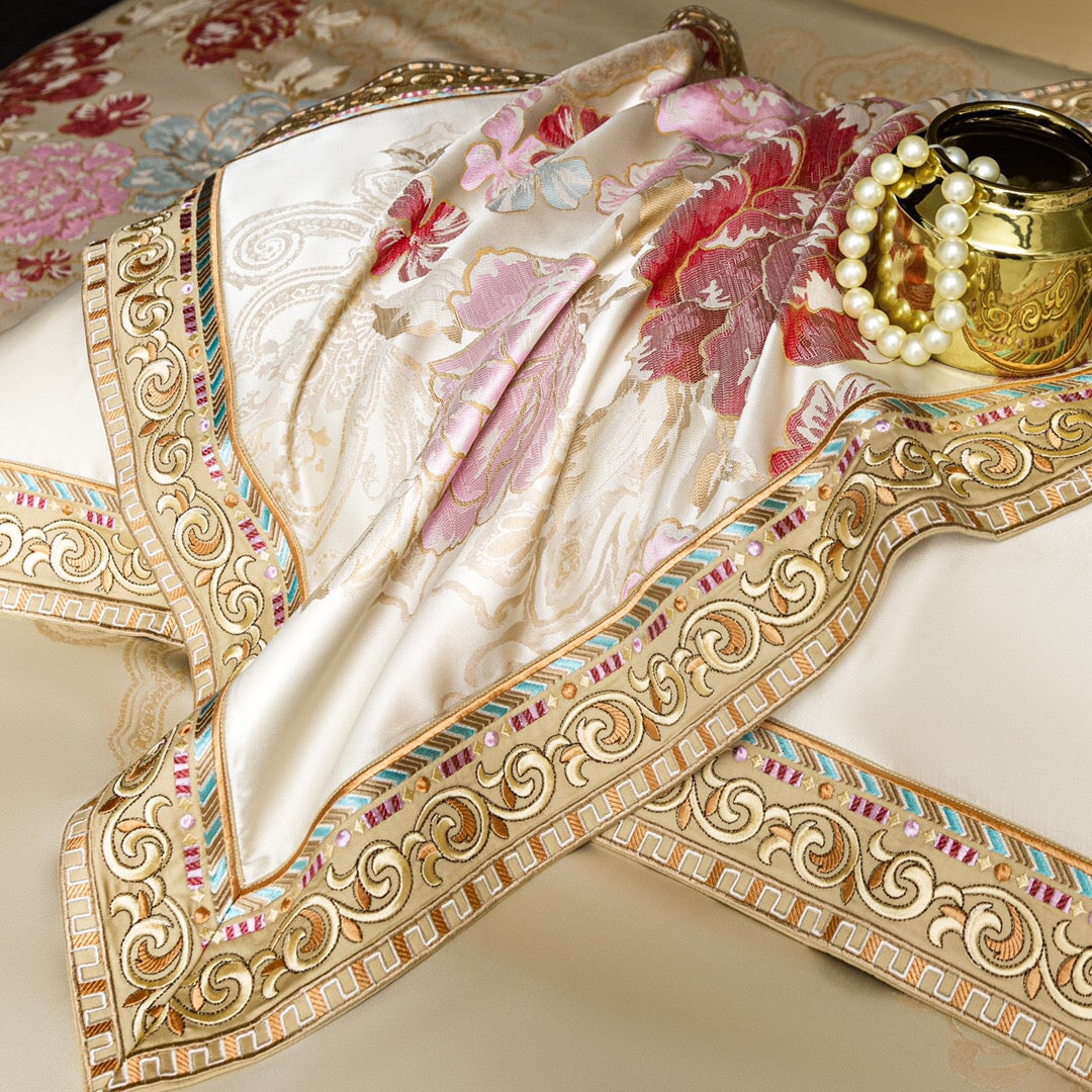 A close-up view of luxurious ivory fabric adorned with intricate floral embroidery in vibrant pink, red, and pastel hues. The edges are framed with an ornate golden border featuring elaborate swirls and colorful accents, including gems and metallic threadwork. A golden decorative vase and a string of pearls are styled alongside, enhancing the opulent and regal aesthetic of the fabric, perfect for premium bedding or home decor.