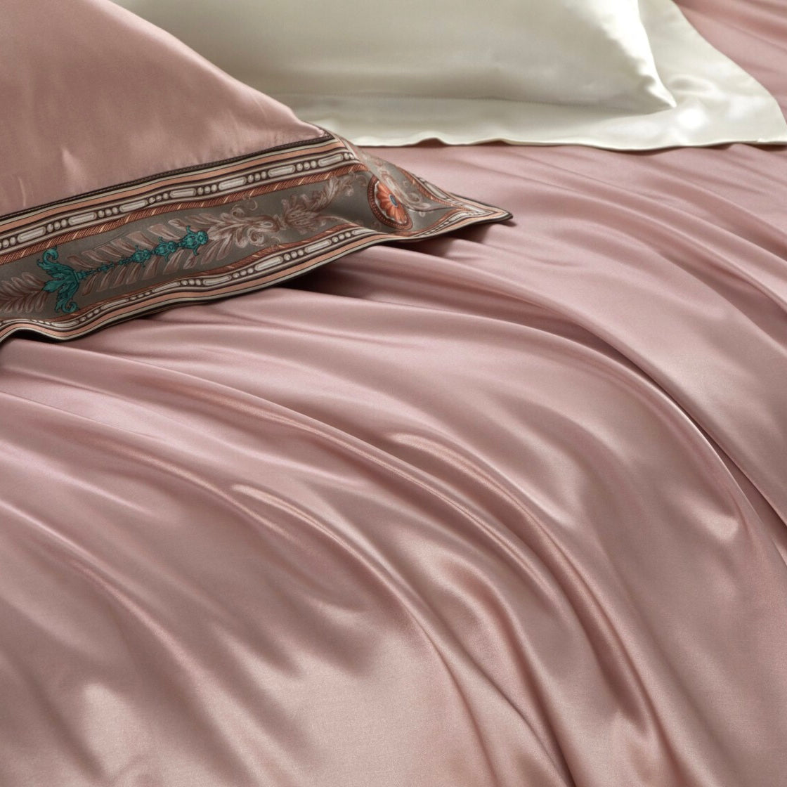 A blush pink bedding set with a smooth, lustrous surface that highlights its luxurious silk texture. The pillowcase features a detailed ornamental border with floral and scrollwork patterns in earthy tones and teal accents, adding a sophisticated touch to the overall elegant design.