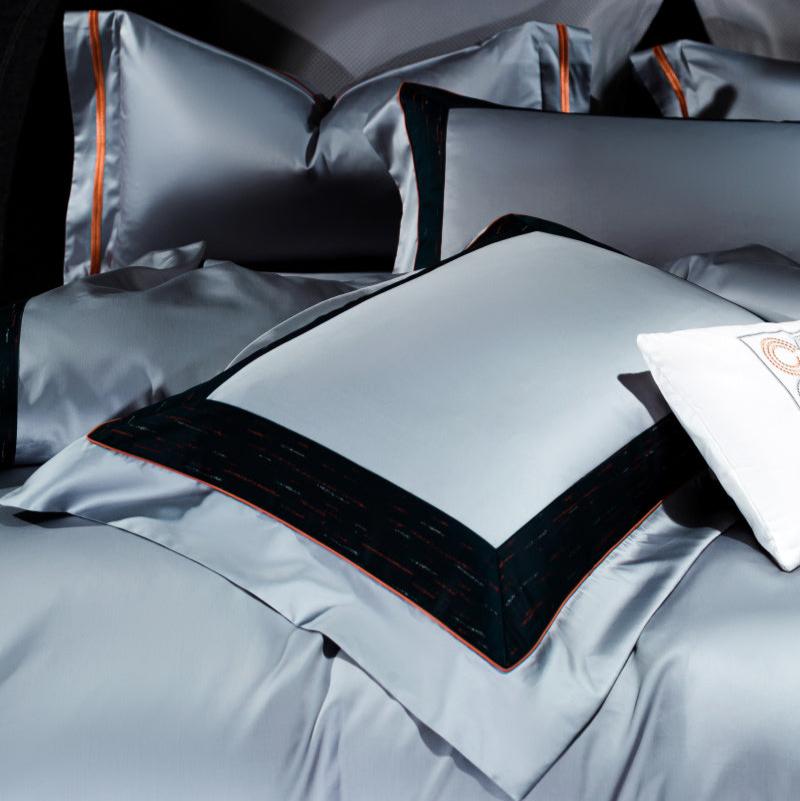 Close-up of luxury bedding set featuring smooth silver fabric with navy blue and red-bordered pillowcases. The sleek design highlights crisp edges and tailored finishes, showcasing a sophisticated and contemporary style. A decorative white pillow with embroidered circular patterns adds contrast to the arrangement.

