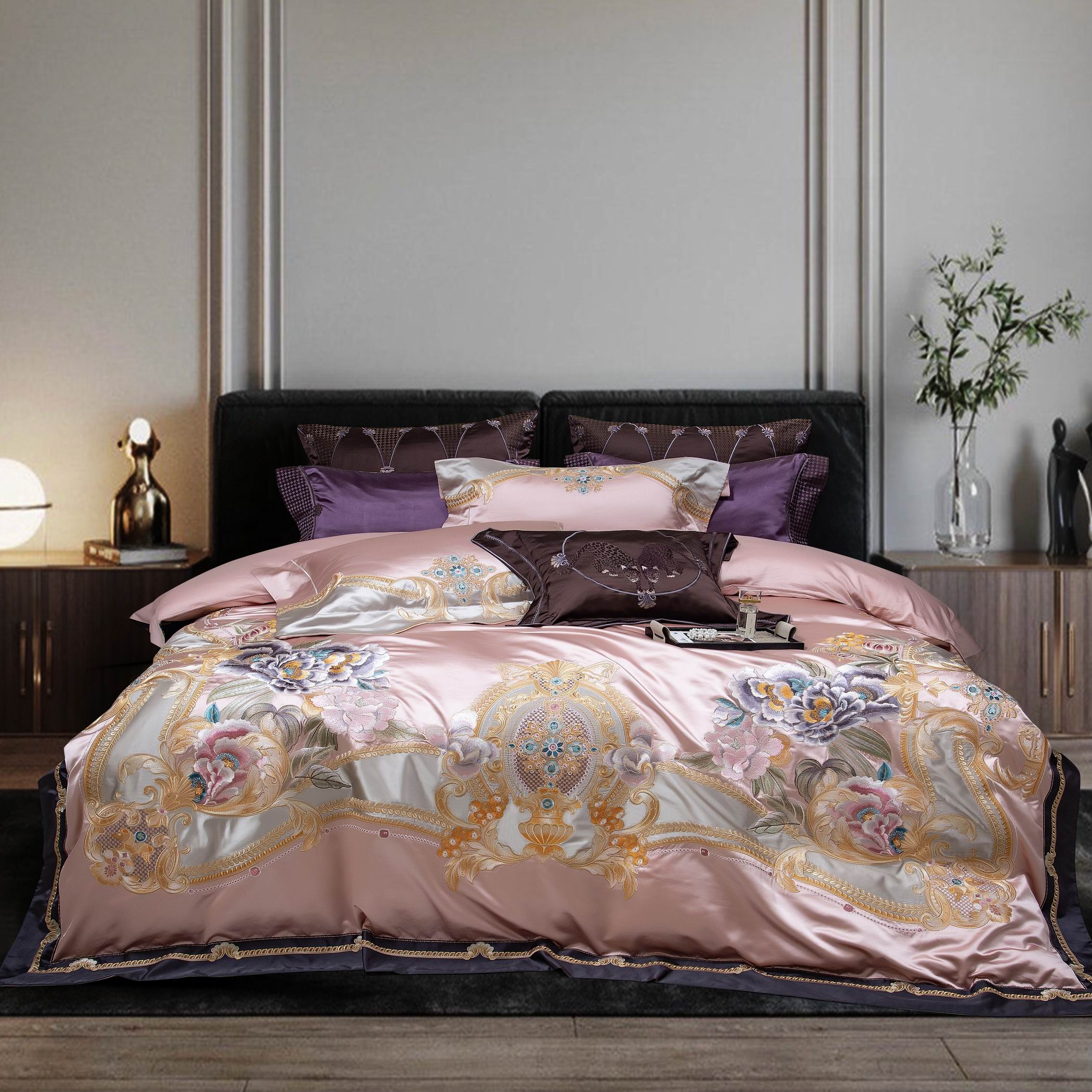 A luxurious bedding set featuring a silky, blush pink duvet adorned with intricate floral embroidery in pastel and gold tones. The design includes ornate patterns and soft contrasts with coordinating dark and light-toned pillows, creating a sophisticated and elegant bedroom ambiance.