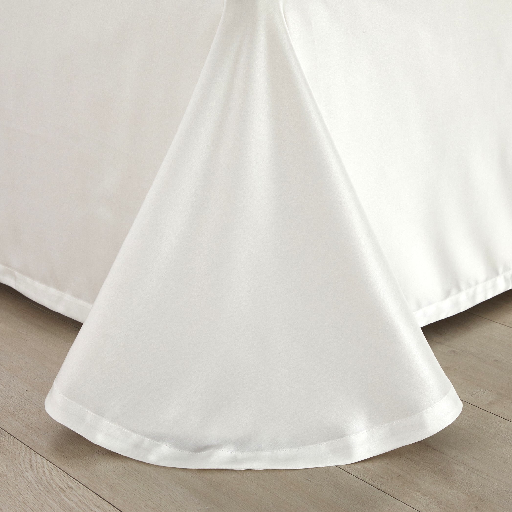 The lower corner of a white bed skirt elegantly draping over a light wood floor, showcasing its smooth texture and precise stitching along the hemline.