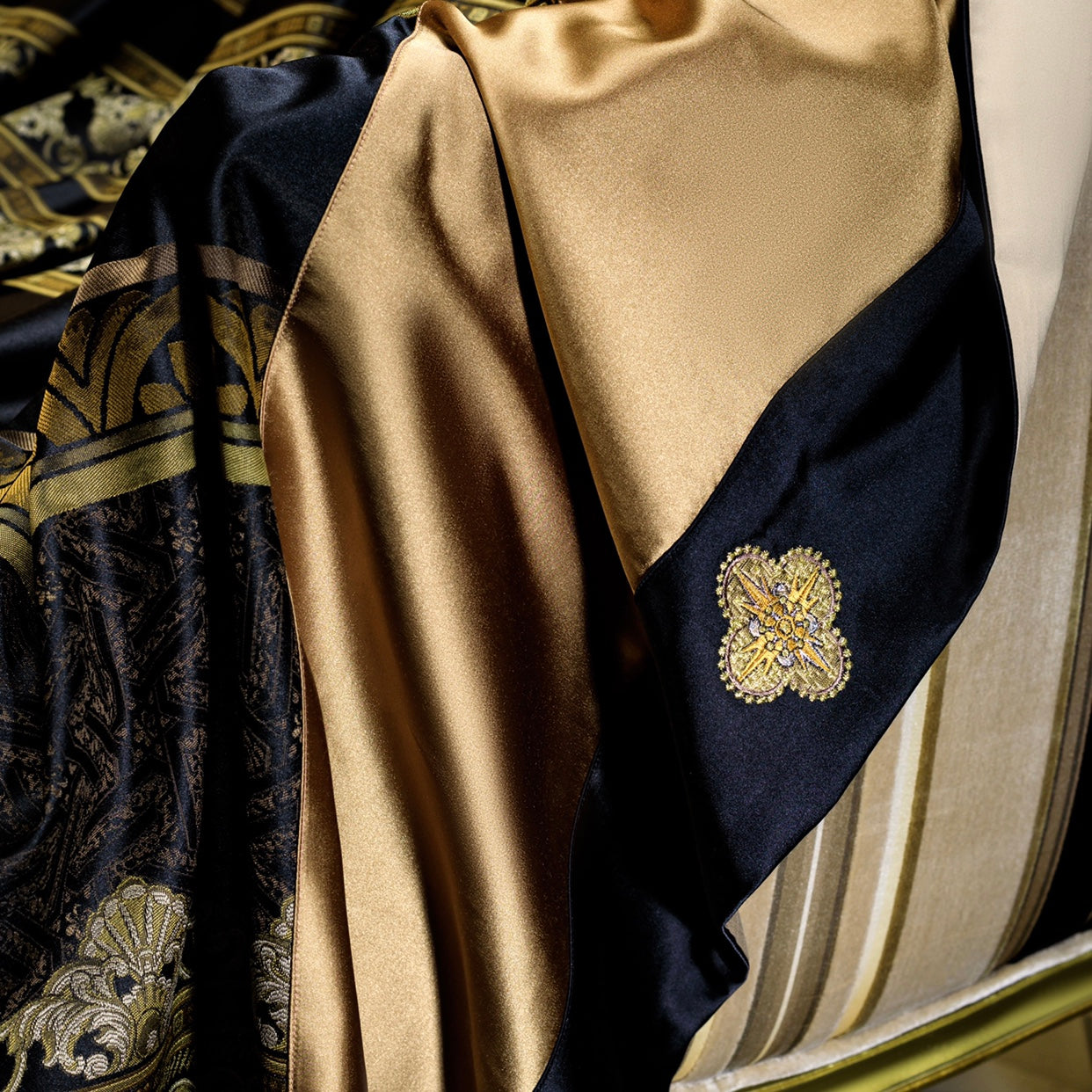 This image highlights a close-up view of a luxurious bedding set's intricate detailing. The fabric showcases a blend of rich black and gold satin, with an ornate embroidered medallion motif adding a regal touch. The smooth and shiny satin texture enhances the opulence, while the corner embroidery reflects fine craftsmanship. The elegant color contrast and detailed embellishment make this bedding piece an ideal choice for an upscale, sophisticated bedroom décor.