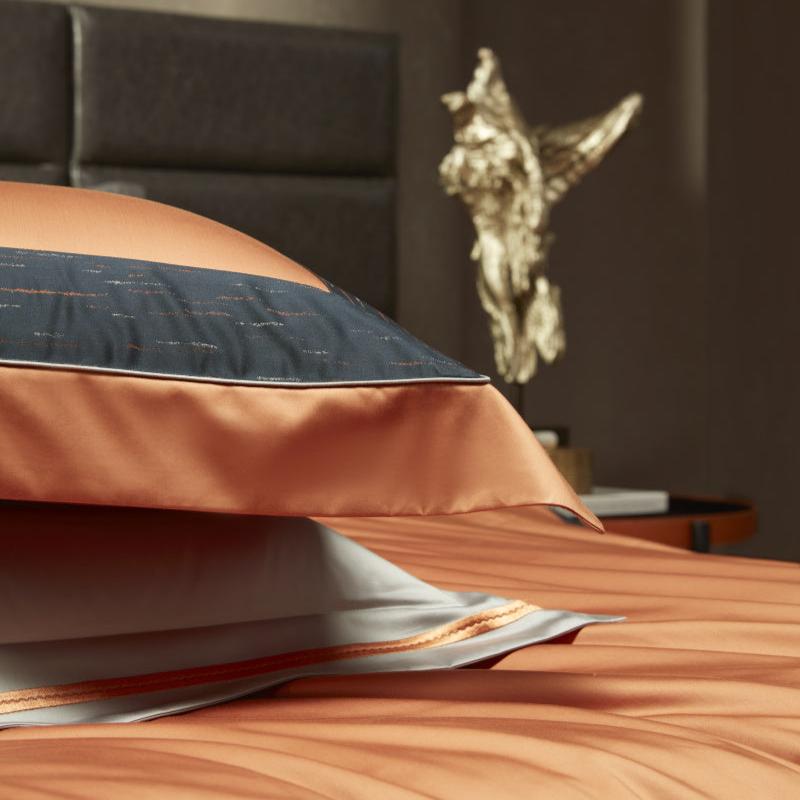 A close-up of stacked pillows and bedding in a warm orange hue, with elegant black fabric trim featuring delicate stitched details. The silky texture and precise piping add a luxurious touch. In the background, a golden sculpture adds an artistic and opulent element to the setting.

