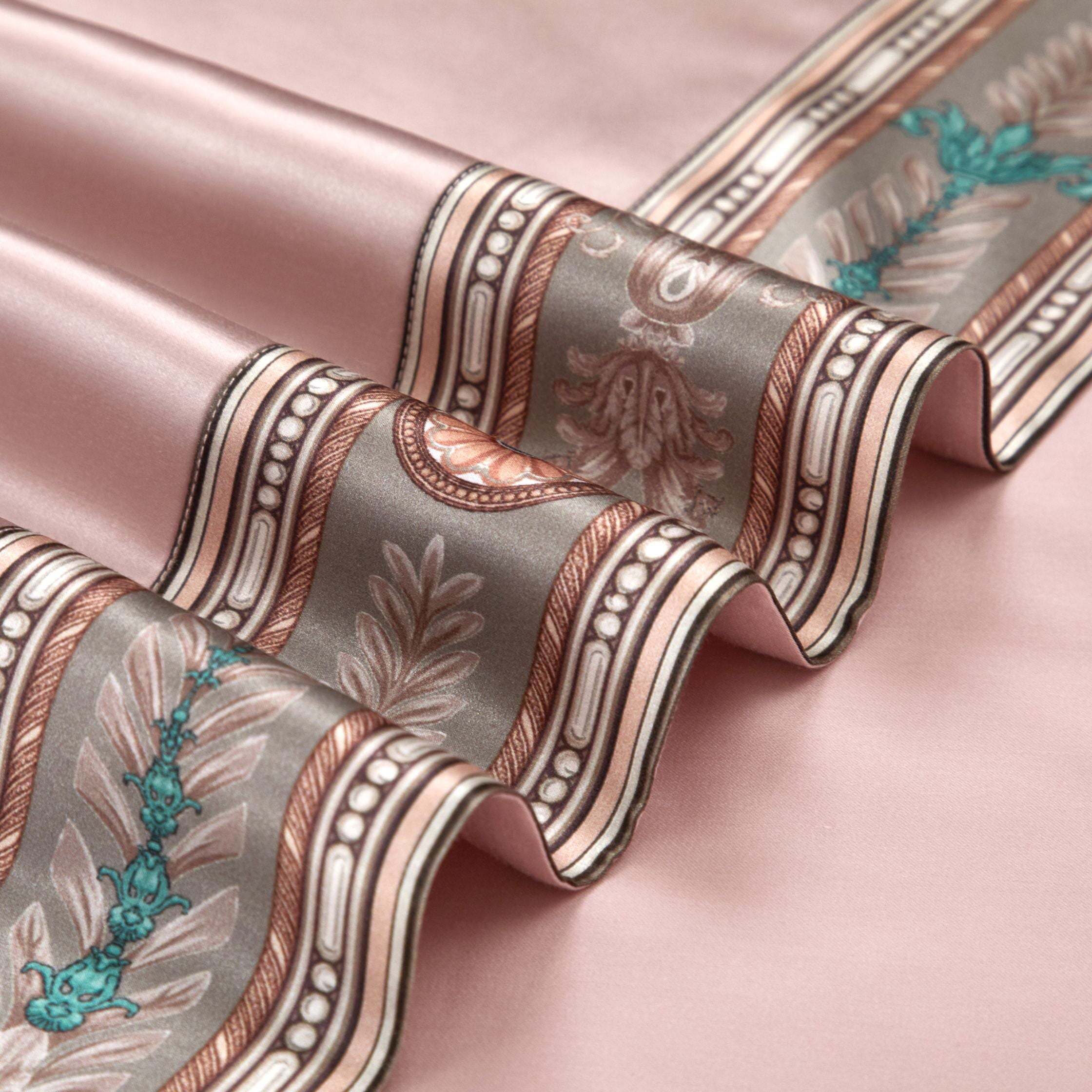 A close-up of the blush pink silk fabric with a prominent ornamental border showcasing intricate patterns. The design includes floral and scrollwork motifs in muted earthy tones, teal accents, and a pearl-like detailing, highlighting the luxurious texture and artistry of the fabric.