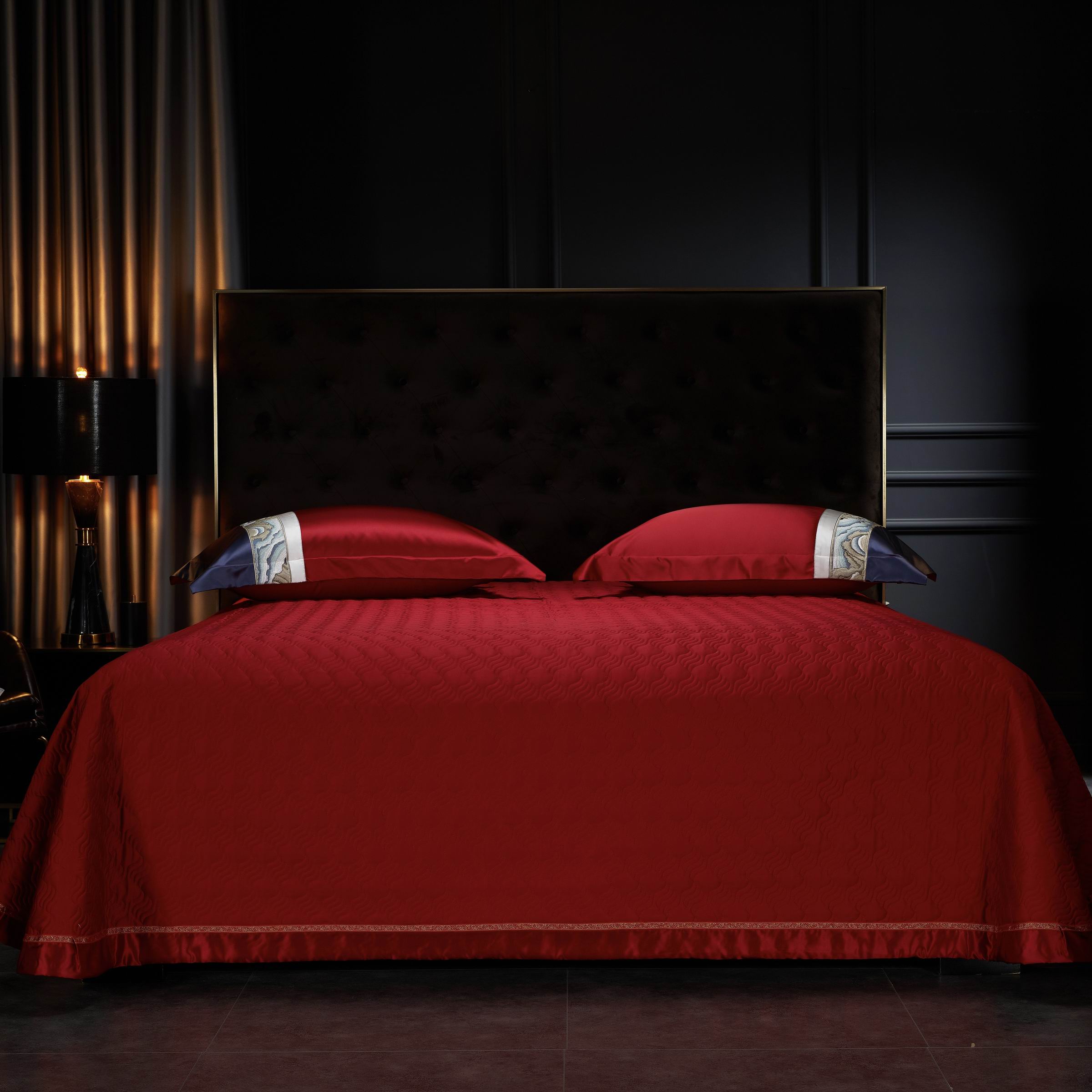 A dark tufted headboard against a black wall, red bedding with a quilted texture, and two red pillowcases featuring a decorative white and navy embroidered trim.