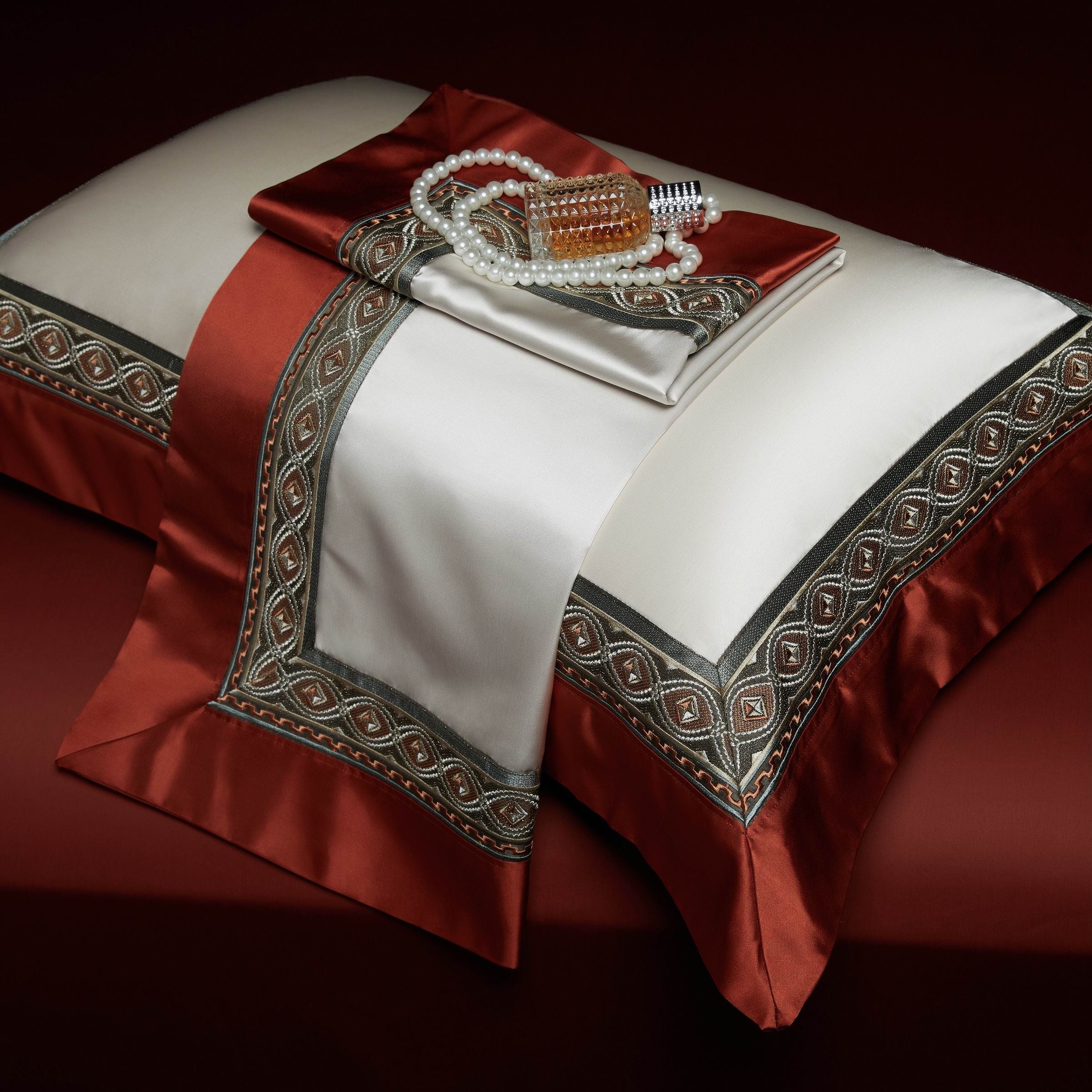 A stunning display of a luxurious pillowcase set in an opulent design. The pillowcases feature a creamy ivory center framed by deep crimson red borders, adorned with intricate olive green and gold embroidery. A matching decorative fabric is elegantly draped across the pillow, complemented by a strand of pearls and a vintage-inspired perfume bottle, evoking a timeless sense of luxury and sophistication. The rich red background enhances the grandeur of the composition.