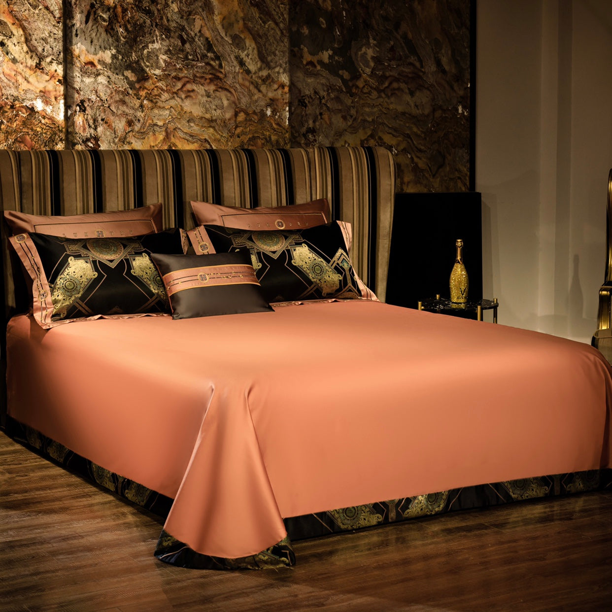 A luxurious bedding setup with a copper-colored flat sheet covering the bed, accented by black pillows with intricate golden geometric designs and copper trim. The striped headboard complements the warm tones, while a marble-textured wall in the background adds an opulent touch. A gold-detailed bottle sits on a side table, enhancing the elegant and sophisticated atmosphere.