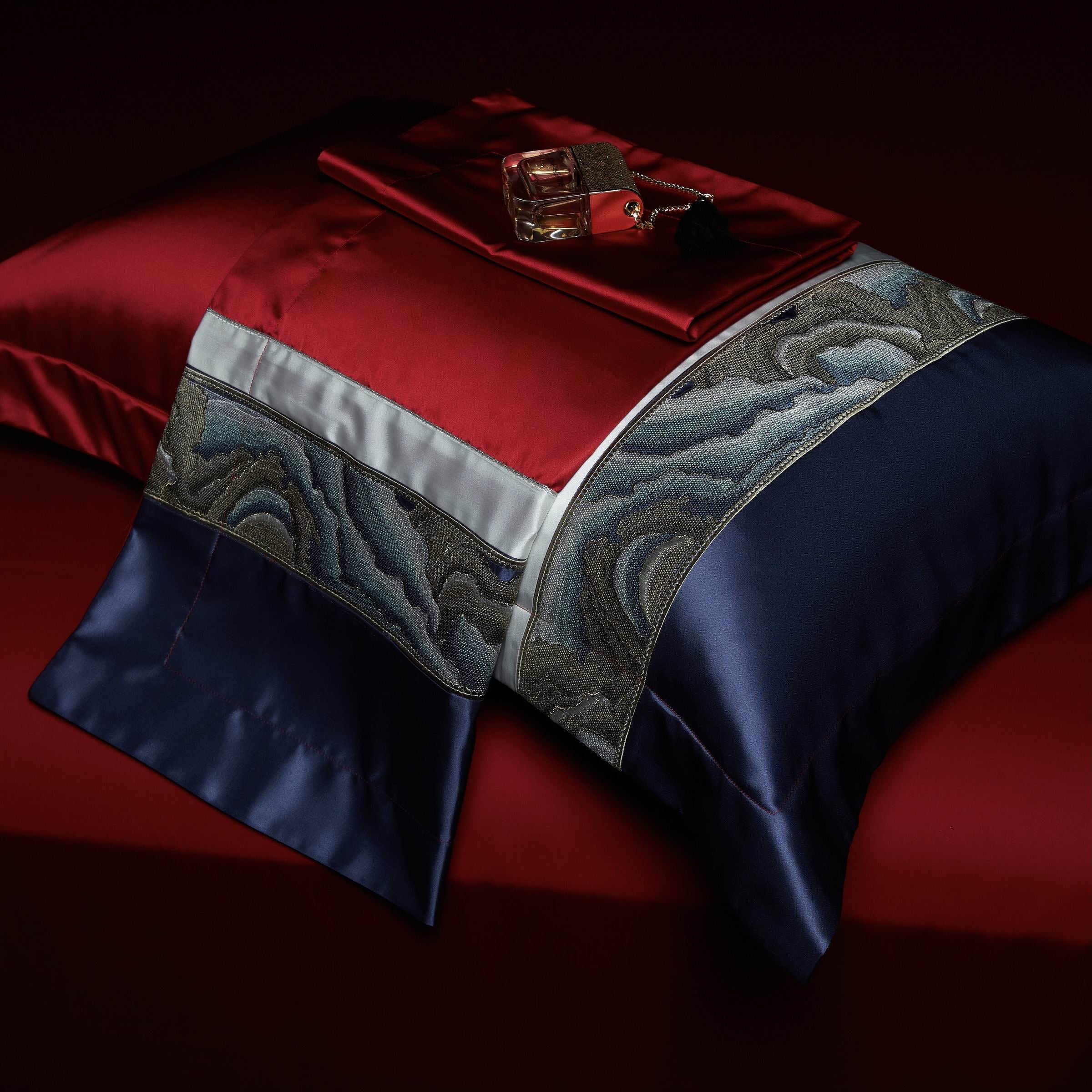 A navy blue and deep red pillowcase with intricate wave-like embroidery on a white trim, accompanied by folded red fabric and a perfume bottle resting on top, set against a dark red background.