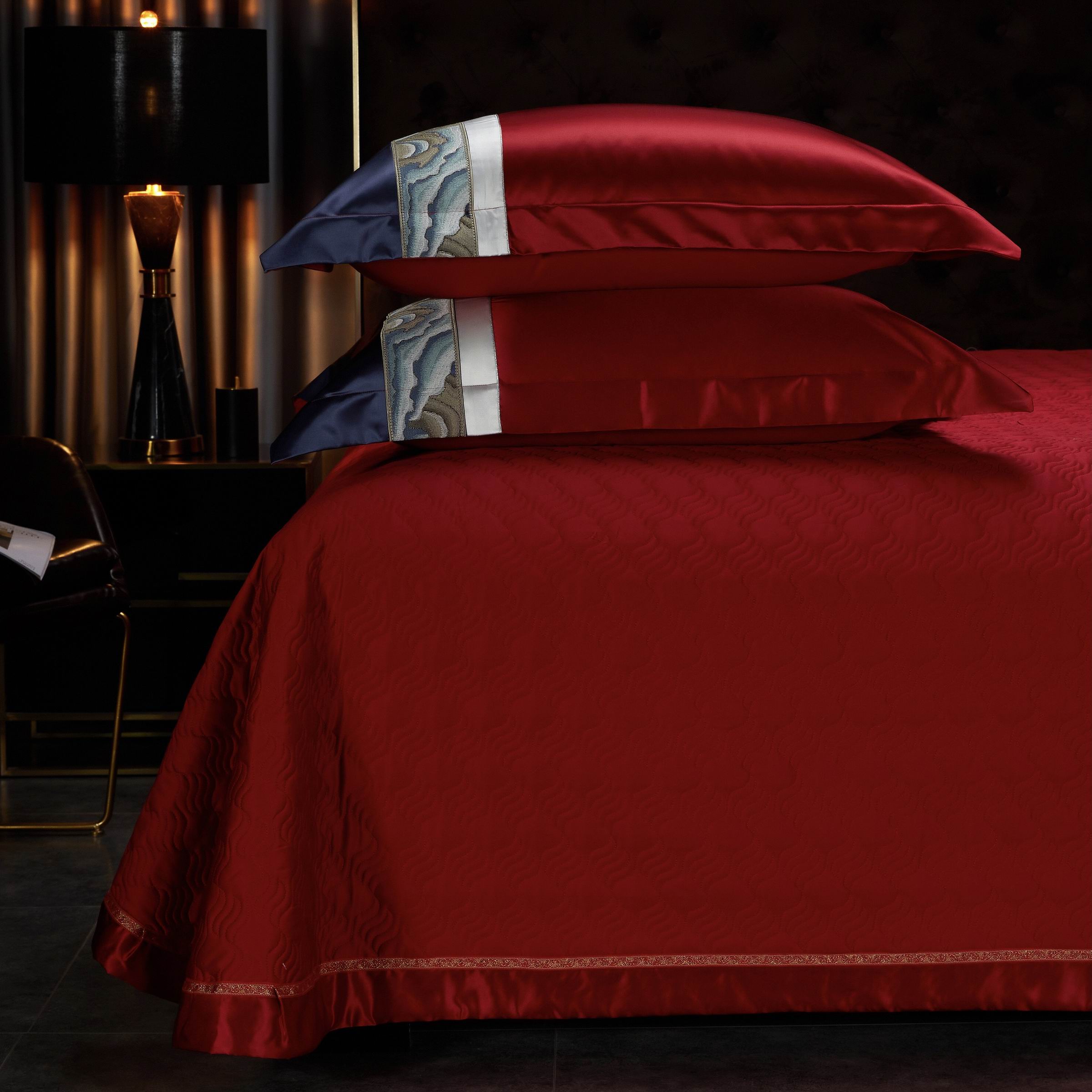 A layered stack of red satin pillows with navy and white embroidered trims featuring intricate wave patterns, placed on a matching red quilted bedspread.