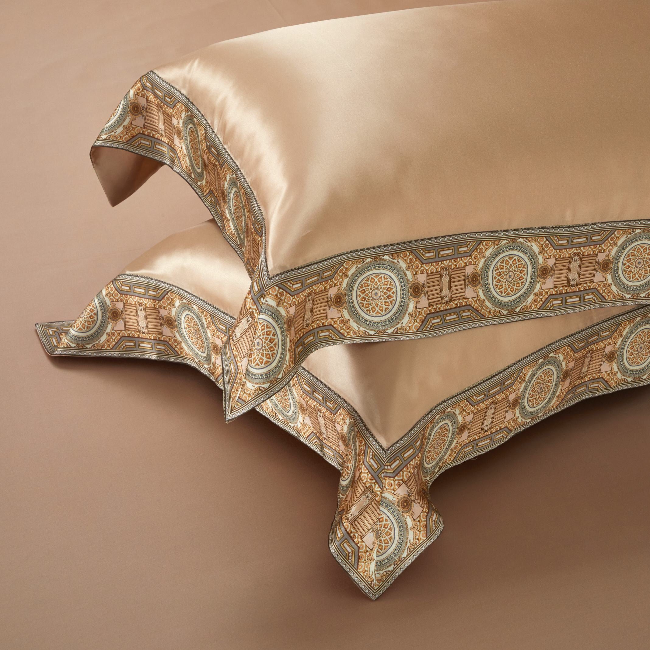 A pair of champagne gold silk pillowcases featuring an intricate border design. The geometric pattern combines warm gold tones with accents of teal and detailed motifs, creating a refined and opulent look. The pillowcases rest on a matching champagne backdrop, emphasizing their elegant craftsmanship and luxurious sheen.